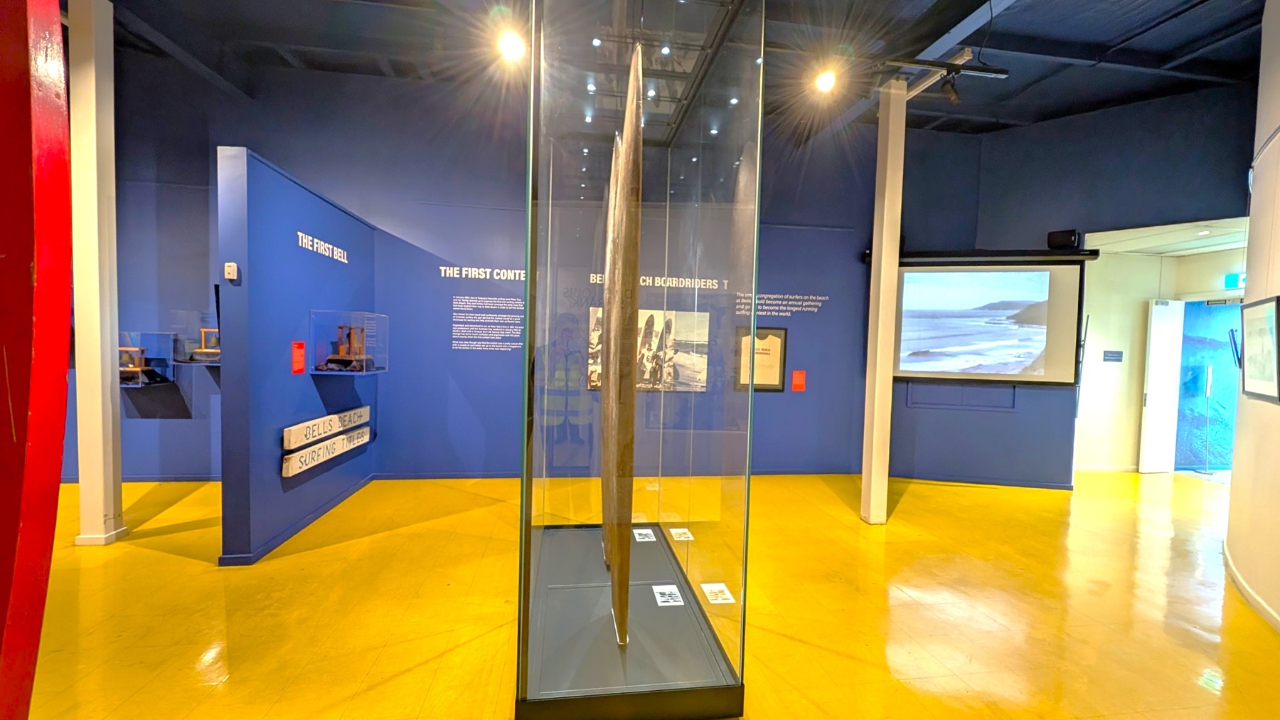 A museum exhibit with a central glass case displaying a long, narrow object. The walls are dark blue with text and images about surfing history. On the left, small display cases are mounted on the wall. On the right, a screen shows a coastal scene. The floor is shiny and yellow, and there are white pillars supporting the ceiling. A doorway is visible in the background.