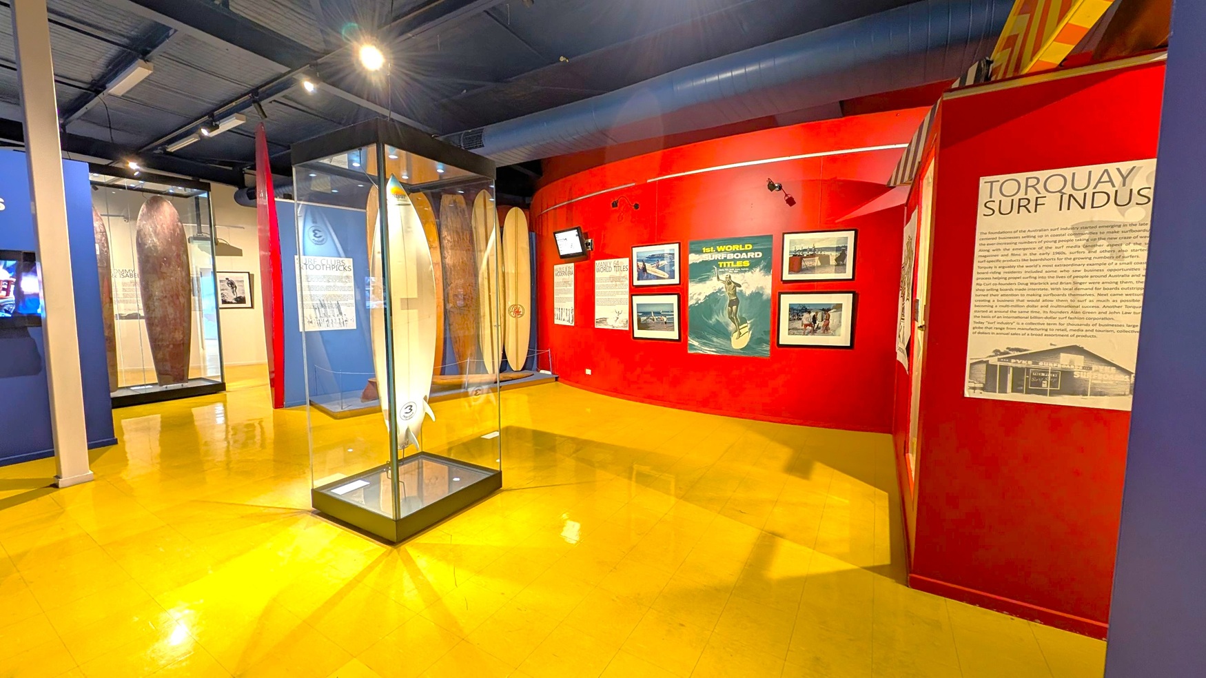 A museum exhibit with a yellow floor and a dark ceiling. In the centre, a glass case displays a white surfboard. The left side features surfboards in glass cases against blue walls. The right side has a red wall with framed photographs and a poster about the "1st World Surfboard Titles." A panel titled "Torquay Surf Indus" provides information. Spotlights illuminate the area.