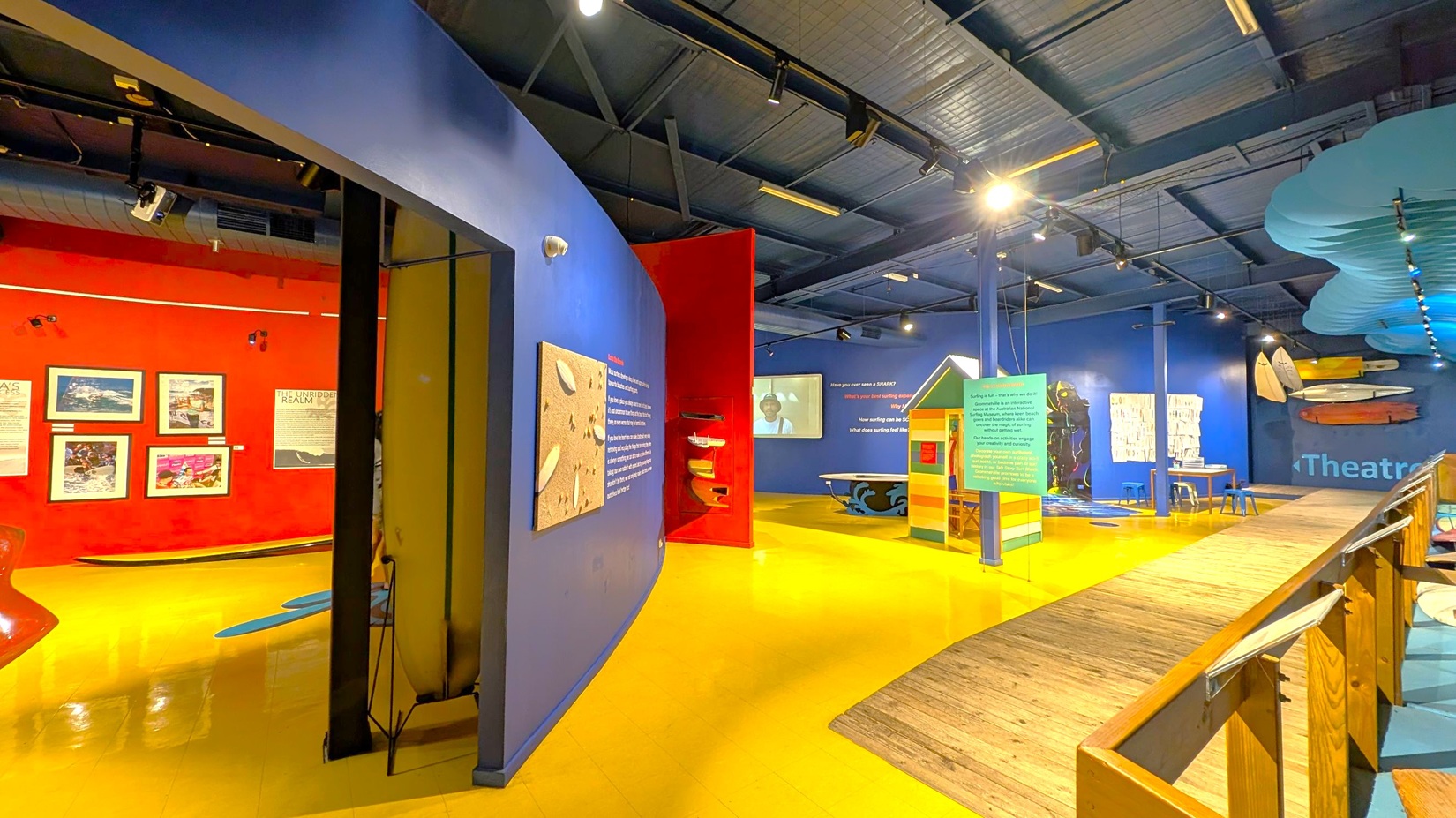 A museum interior with vibrant colours. On the left, a red wall displays framed pictures and surfboards. The floor is bright yellow. In the centre, a blue wall features a textured artwork and text. A small, colourful beach hut stands nearby. On the right, a blue wall with surfboards and a sign reading "Theatre" is visible. The ceiling is dark with exposed beams and spotlights. A wooden walkway runs along the right side.
