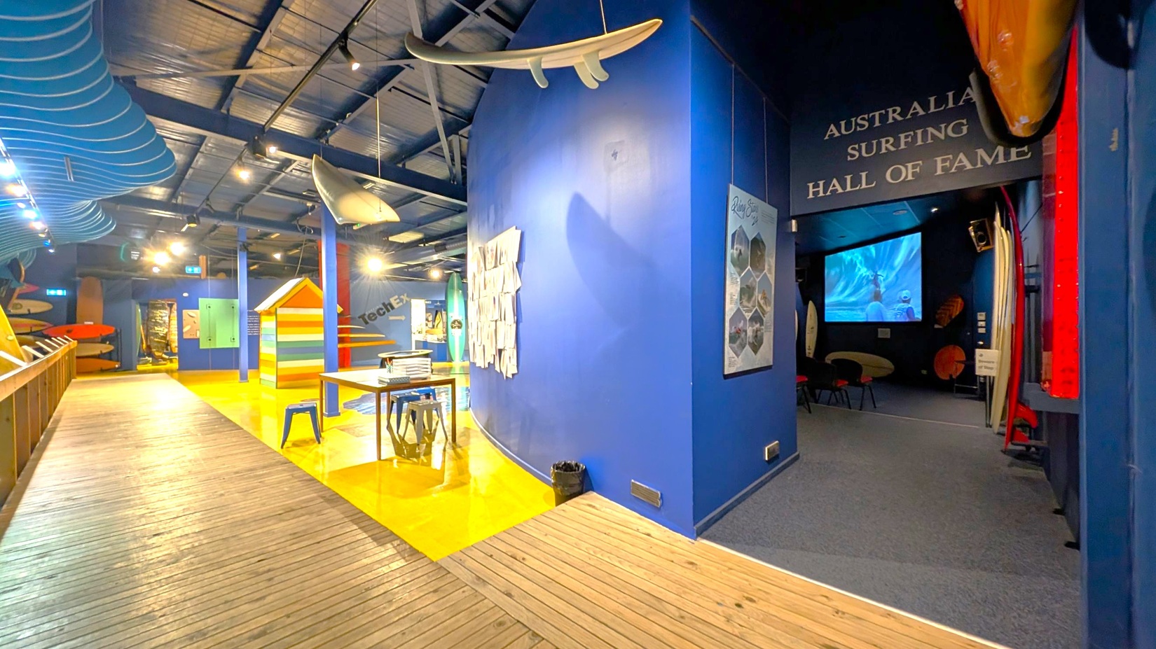 A museum interior with a wooden floor and a high, dark ceiling. On the left, surfboards are displayed along the wall. In the centre, a colourful beach hut and a table with stools are visible. A surfboard hangs from the ceiling. On the right, a darkened area with a screen showing a surfing video is labelled "Australian Surfing Hall of Fame." The walls are painted blue, and there are informational posters and displays throughout.