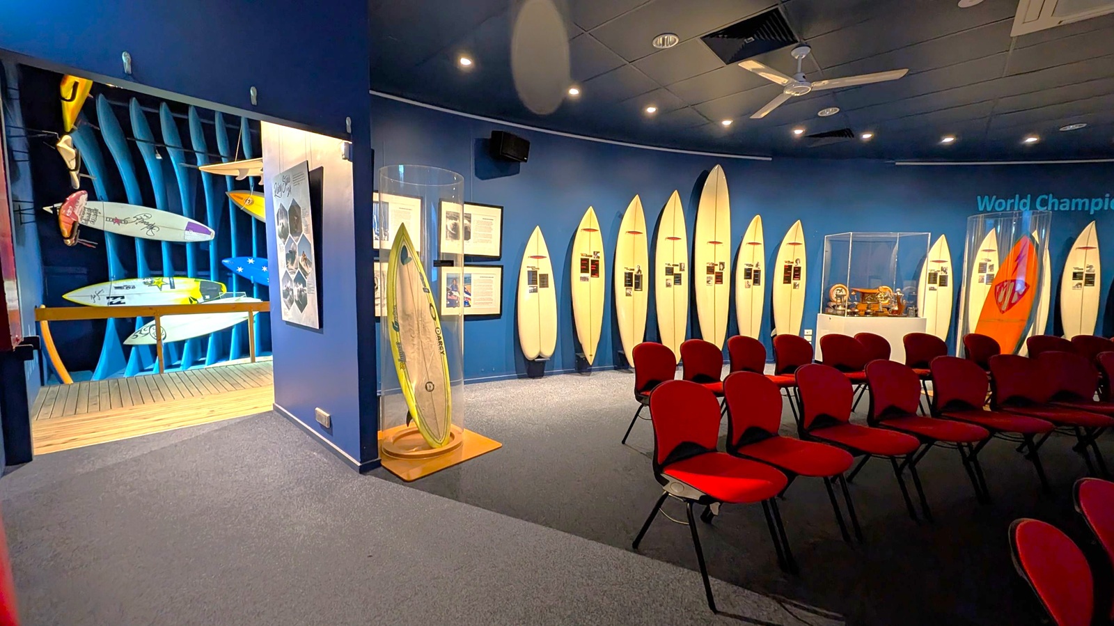 A room with a display of surfboards on the right wall, each accompanied by informational plaques. In the foreground, there are rows of red chairs. On the left, an open doorway reveals more surfboards mounted on a blue wall with a wooden floor. The ceiling is dark with spotlights and a ceiling fan. A glass case with a surfboard stands near the entrance.