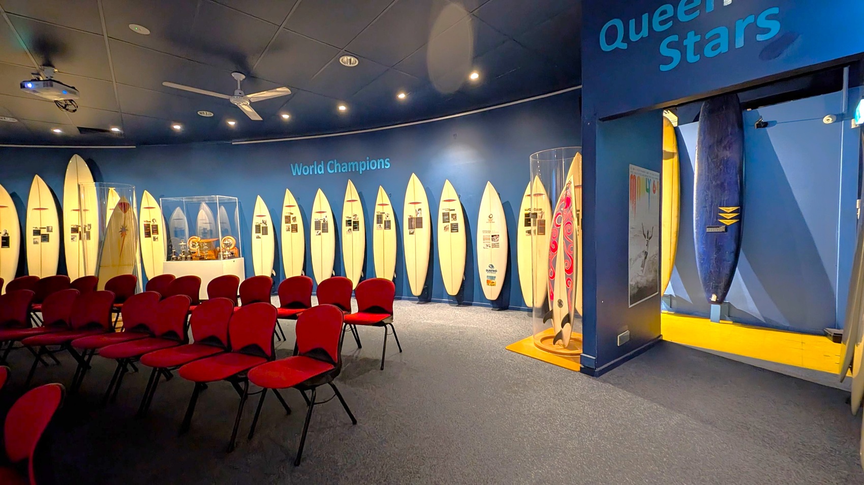 A museum exhibit featuring a row of surfboards displayed vertically along a curved blue wall with the words "World Champions" above. Each surfboard has accompanying information. In the foreground, there are rows of red chairs. To the right, a glass case contains a surfboard with a red swirl design. A doorway leads to another area with a blue surfboard and a poster. The ceiling is dark with spotlights and a ceiling fan.