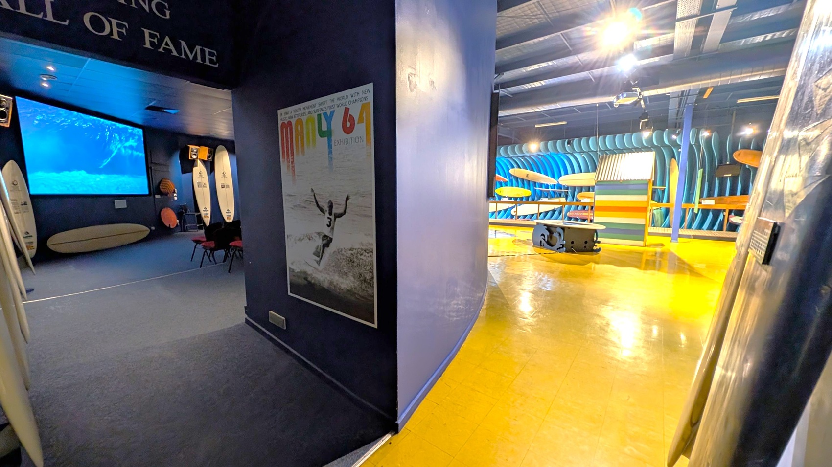 On the left, a dark room with a large screen showing an underwater scene, surrounded by surfboards and chairs. A poster is on the wall near the entrance. On the right, a brightly lit area with a yellow floor, displaying multiple surfboards against a blue, wave-like backdrop. The ceiling has exposed beams and lighting fixtures.