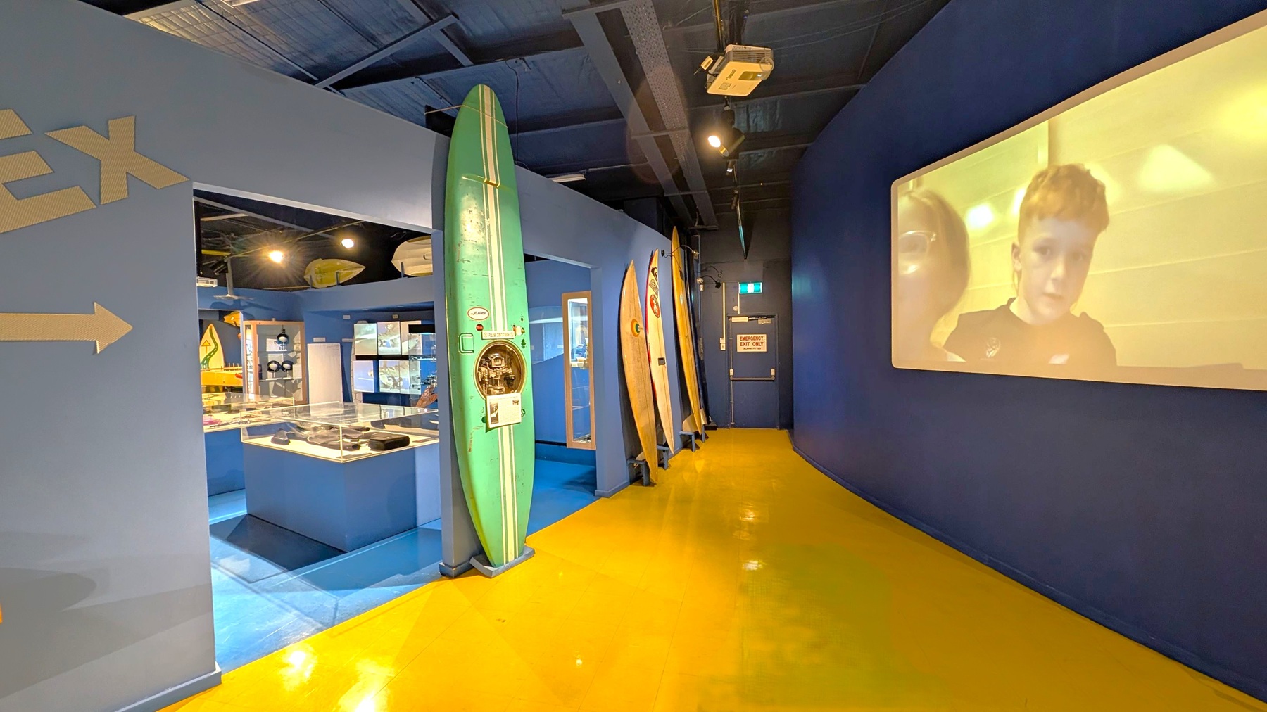 A museum exhibit with a blue and yellow colour scheme. On the left, there are surfboards displayed vertically against a blue wall. A glass display case with various items is visible further inside. On the right, a large screen shows an image of two people. The floor is bright yellow, and the ceiling is dark with exposed fixtures. An emergency exit door is at the end of the corridor.