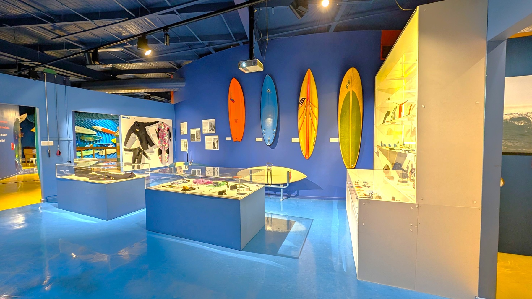 A museum exhibit with a blue floor and ceiling. On the left, there are display cases with various surfing equipment and wetsuits. The back wall features three surfboards in orange, blue, and yellow, mounted vertically. To the right, a large glass cabinet displays smaller surfing-related items. The walls are painted blue, and there are informational panels and photographs throughout the space.