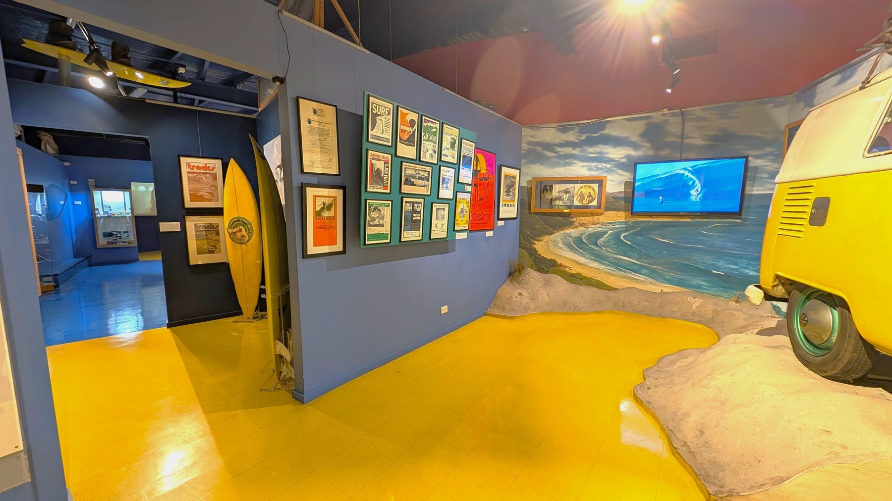 A museum exhibit with a surf theme. On the left, there are framed posters and a yellow surfboard against a dark wall. The floor is yellow. In the middle, a wall displays various framed surf-related posters. On the right, a mural of a beach scene with a screen showing a surfing video. The front of a yellow van is partially visible, positioned on a sandy display. The ceiling is dark with spotlights.