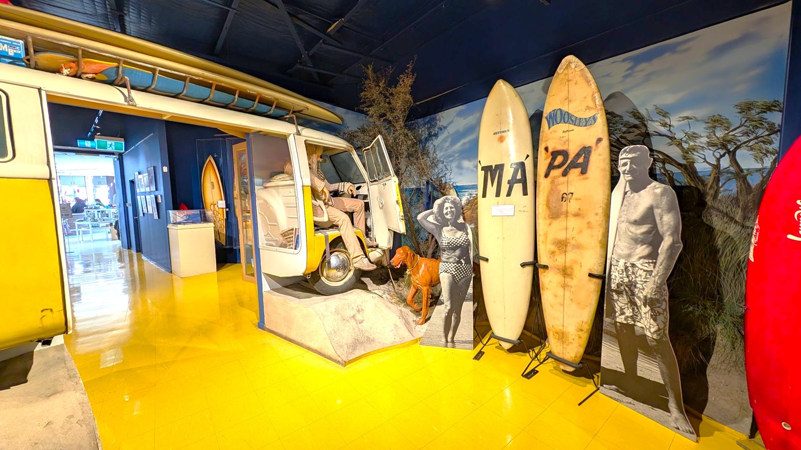 A museum exhibit featuring a yellow and white vintage van with surfboards on the roof. Inside the van, a mannequin is seated. To the right, there are several surfboards standing upright, with two black-and-white cutouts of people in swimwear. The background has a mural of a beach scene with trees. The floor is shiny and yellow, and there is an open doorway leading to another room with tables and chairs visible.