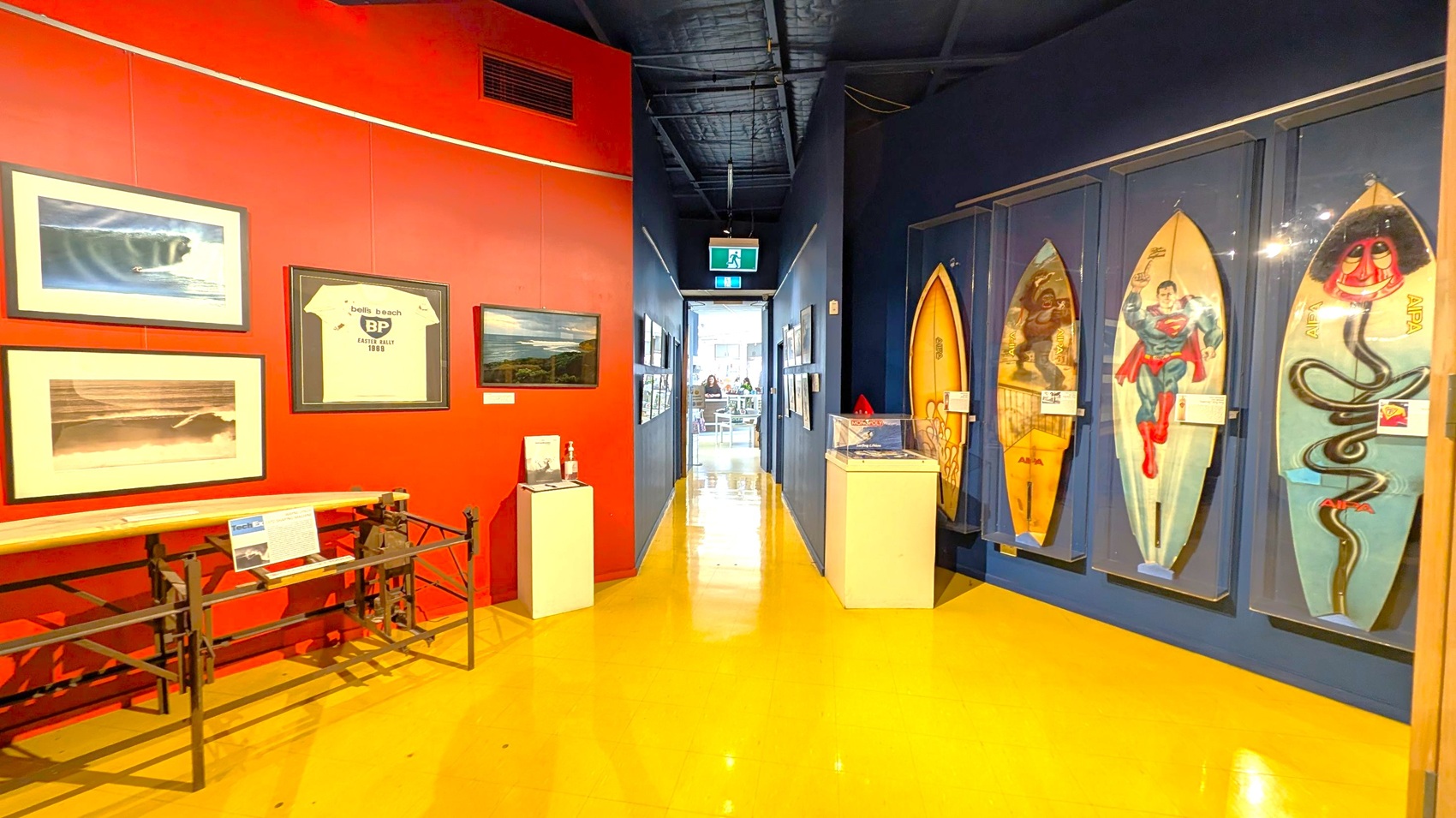 A hallway with a bright yellow floor and a dark ceiling. On the left, a red wall displays framed pictures and a framed T-shirt. Below, a surfboard is mounted on a metal stand. On the right, a blue wall showcases several surfboards with various designs, including a Superman graphic. In the centre, a narrow passage leads to a well-lit room with visible tables and chairs. An exit sign hangs above the passage.