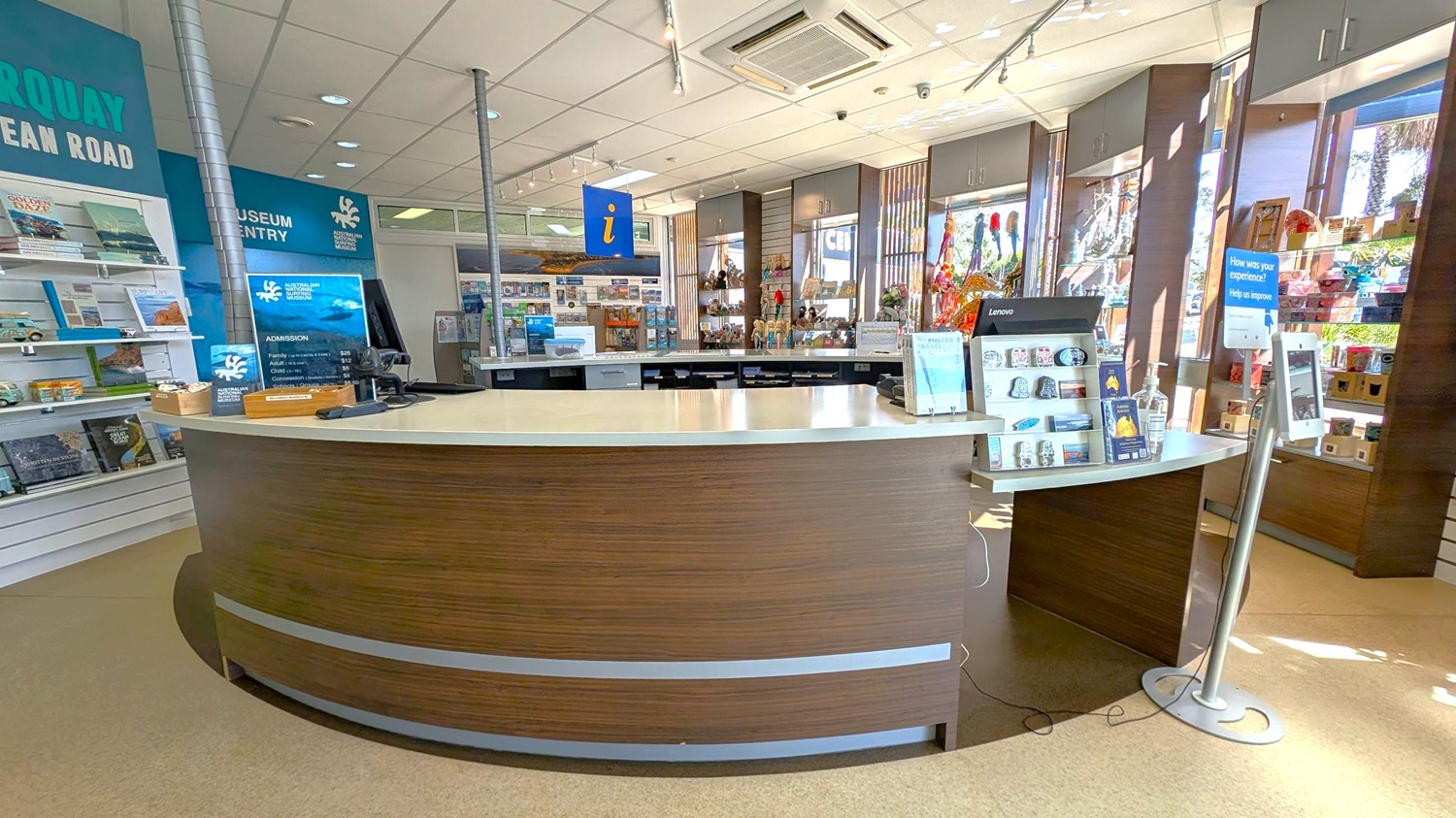 A visitor information centre with a curved wooden reception desk in the foreground. On the left, shelves display brochures and books. Behind the desk, a blue sign with an "i" hangs from the ceiling. The background features large windows with sunlight streaming in, showcasing various souvenirs and gifts. The ceiling is fitted with recessed lights and an air conditioning unit. A digital kiosk stands to the right of the desk.
