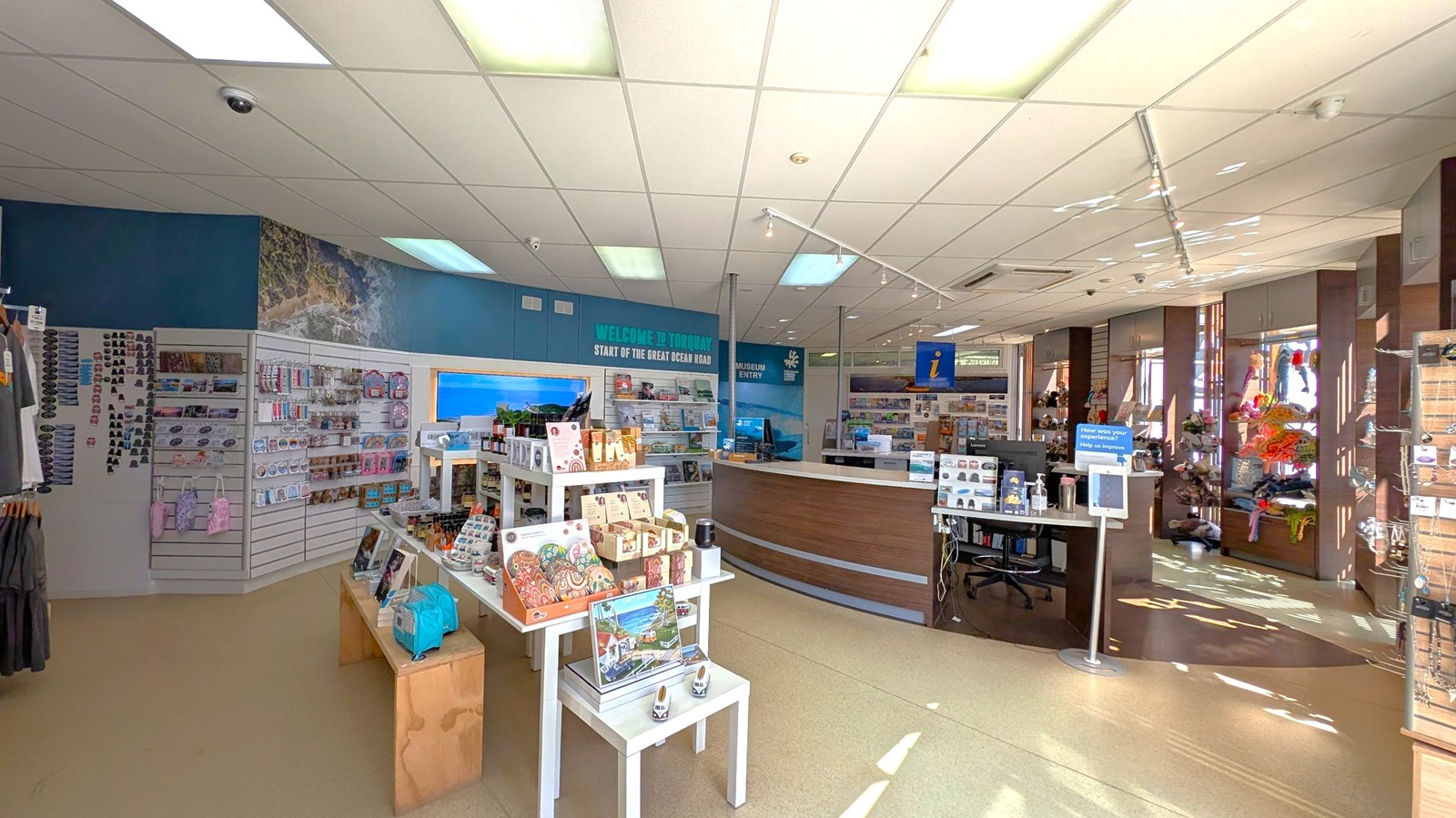 A tourist information centre with a variety of merchandise on display. On the left, shelves hold souvenirs like magnets and keychains. In the centre, white tables showcase local products and postcards. The right side features a curved reception desk with brochures and a computer. Large windows allow natural light to illuminate the space. The ceiling is lined with fluorescent lights, and the floor is tiled in a light colour. Signs indicate the start of the Great Ocean Road and museum entry.