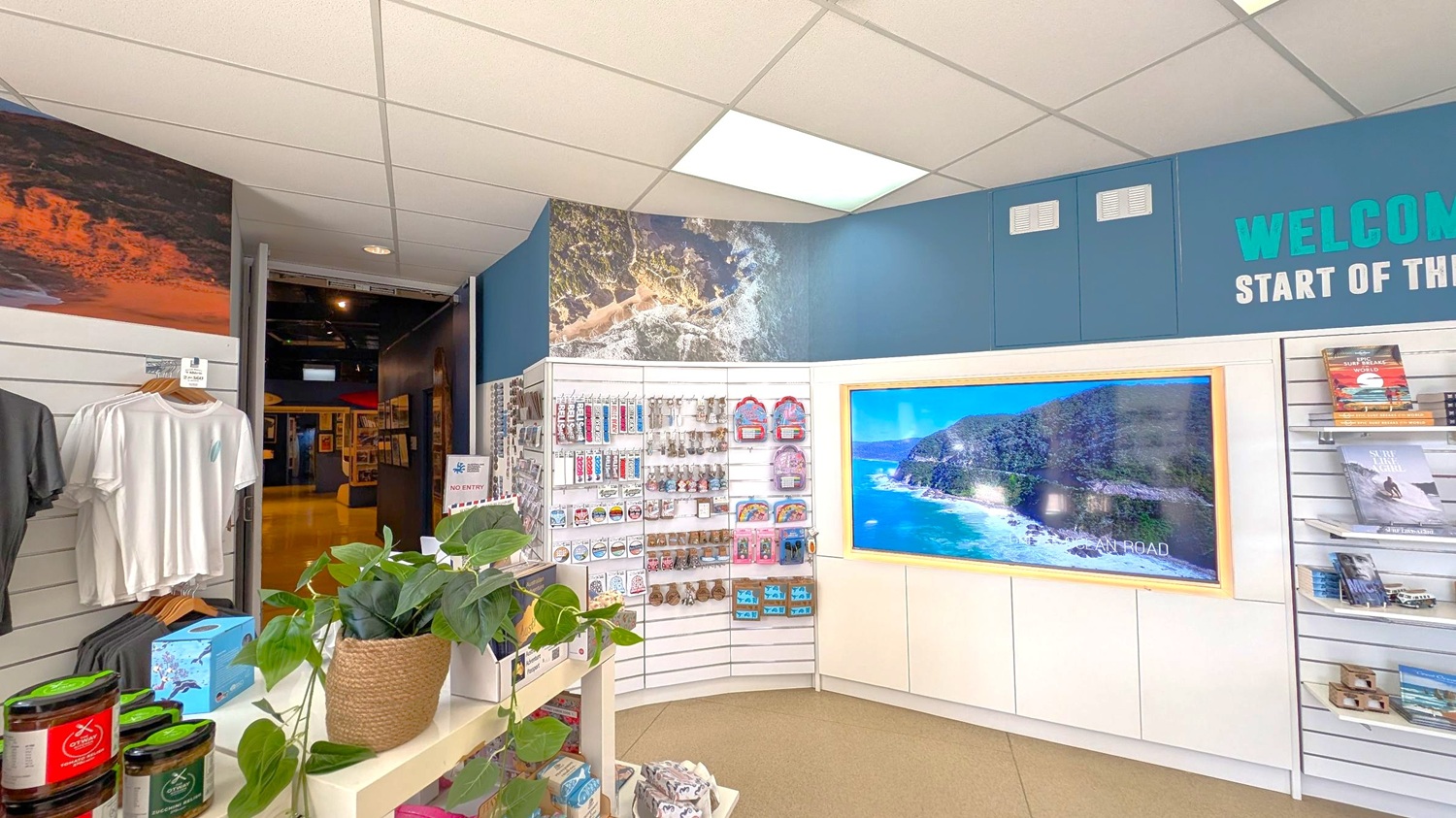 A shop interior with a curved display wall on the right featuring a large screen showing a coastal scene. Shelves hold various souvenirs, including keychains and small bags. On the left, there are hanging T-shirts and a shelf with jars. A potted plant sits on a counter in the foreground. The ceiling is white with fluorescent lights, and the floor is carpeted. A sign above reads "WELCOME START OF THE" with more text obscured.