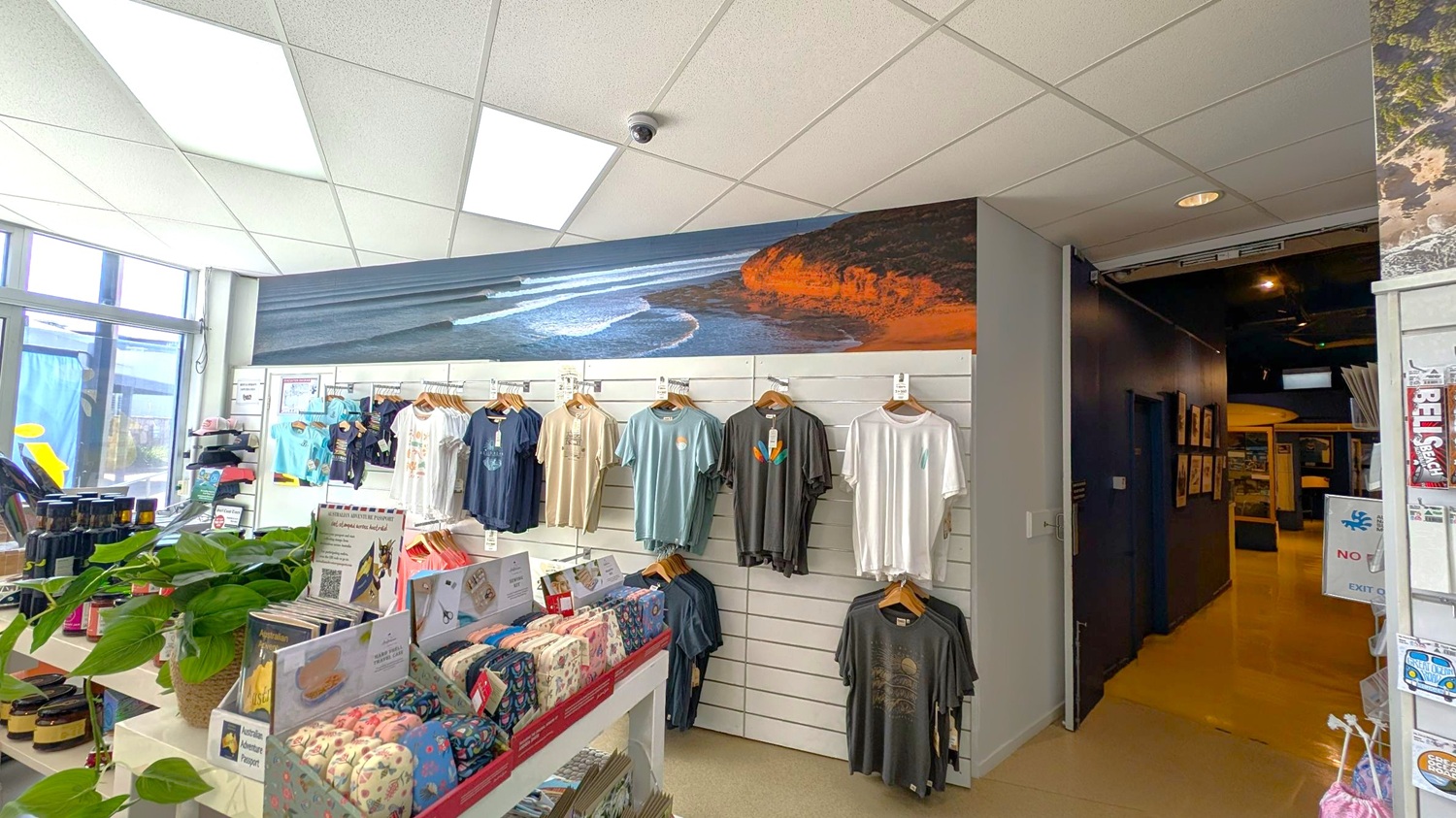 A shop interior with a display of T-shirts hanging on a white slatted wall in the centre. Above the shirts is a large mural of a coastal scene with waves and cliffs. On the left, there are shelves with various products, including jars and a potted plant. The ceiling is made of white tiles with fluorescent lights. To the right, there is a dark hallway leading further into the store. The floor is light-coloured.