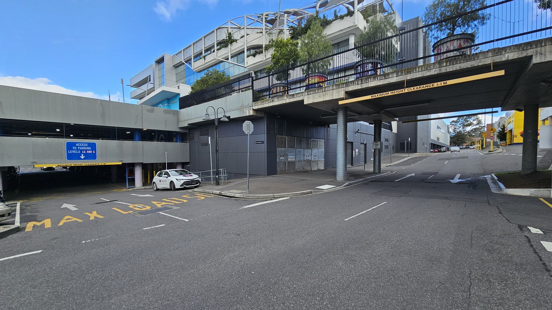 A multi-level car park entrance is on the left, with a sign indicating access to parking levels LG and G. A white car is parked near the entrance. The road has markings for maximum load and directional arrows. On the right, a road passes under a bridge with a clearance sign. Above, there are buildings with modern architecture and greenery. The pavement is visible, and there are street lamps and signs along the road.