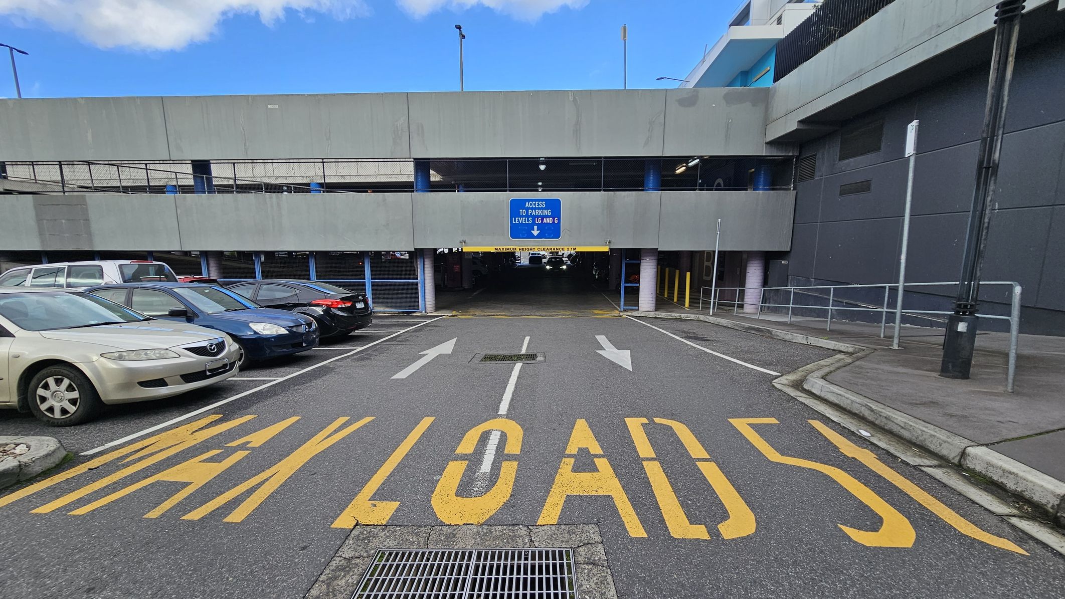 A multi-storey car park entrance with a sign indicating access to parking levels LG and G. The ground has large yellow text reading "MAX LOAD 5T" and directional arrows. On the left, several parked cars are visible, including a beige sedan. On the right, a pavement with a railing and a tall lamp post. The building is grey with a blue sign, and the ceiling of the entrance is low with a clearance warning.