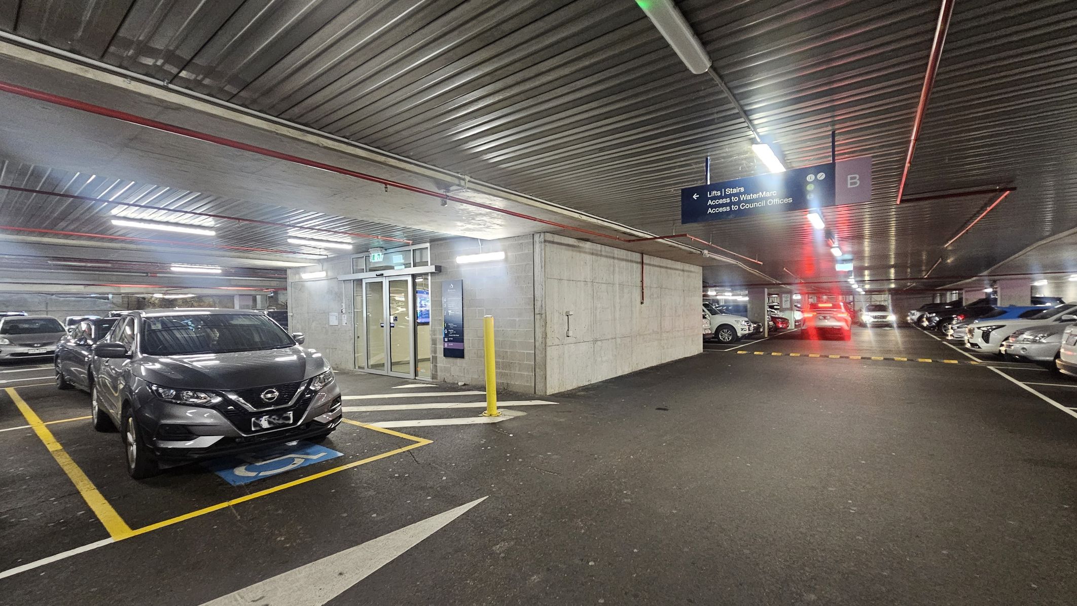 A multi-storey car park with a grey car parked in a disabled parking space on the left. The floor is marked with yellow lines and a blue disabled symbol. In the centre, there is a concrete wall with glass doors leading to lifts and stairs. A yellow bollard is in front of the entrance. On the right, more cars are parked in marked spaces. The ceiling is metallic with fluorescent lights and red pipes. A sign above indicates directions to lifts, WaterMarc, and council offices.