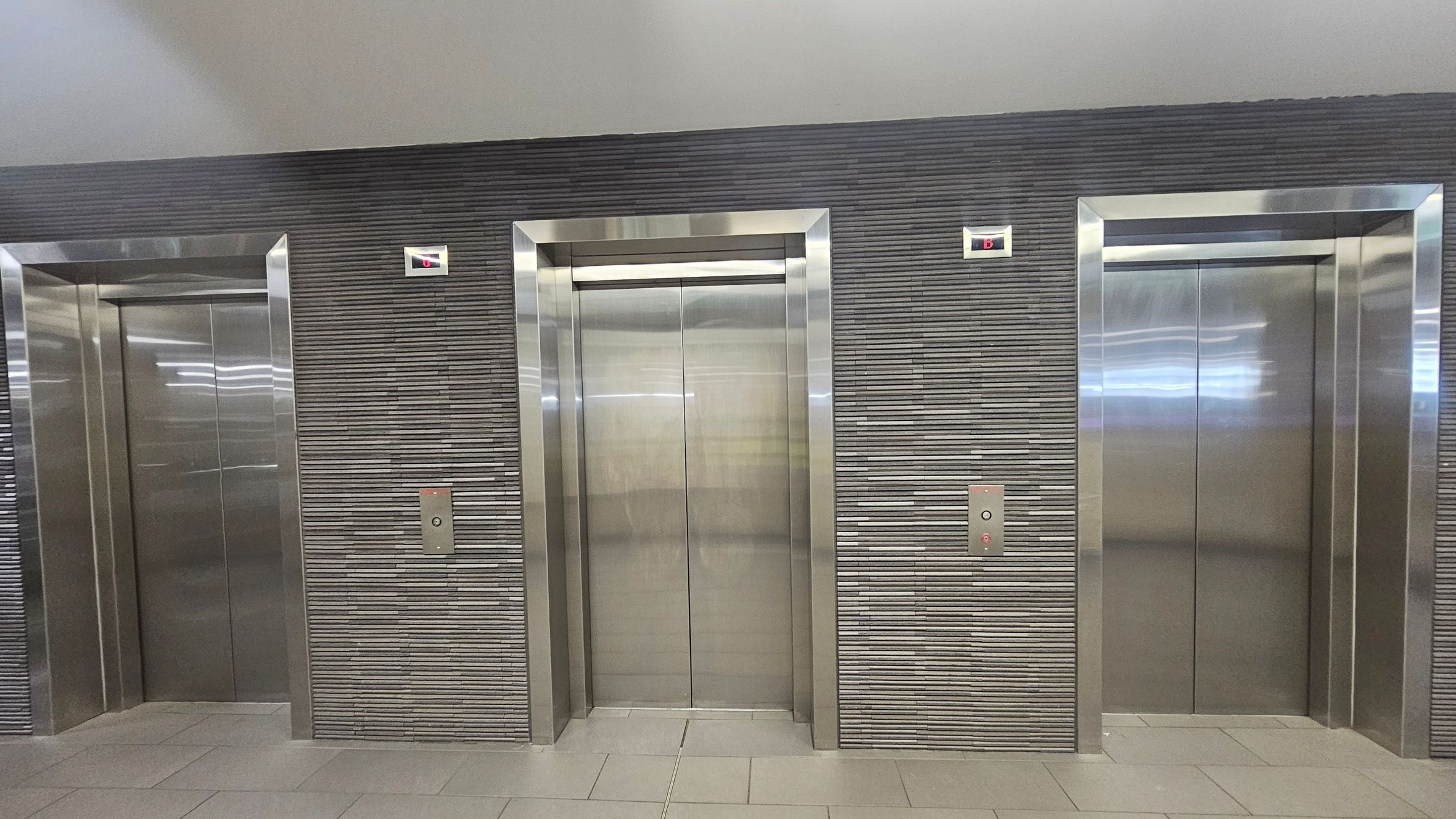 Three stainless steel lift doors are set into a wall with horizontal grey tiles. Each lift has a control panel with buttons and a digital display above showing the letter "B". The floor is tiled in a light grey, and the ceiling is smooth and light-coloured. The lifts are evenly spaced along the wall.
