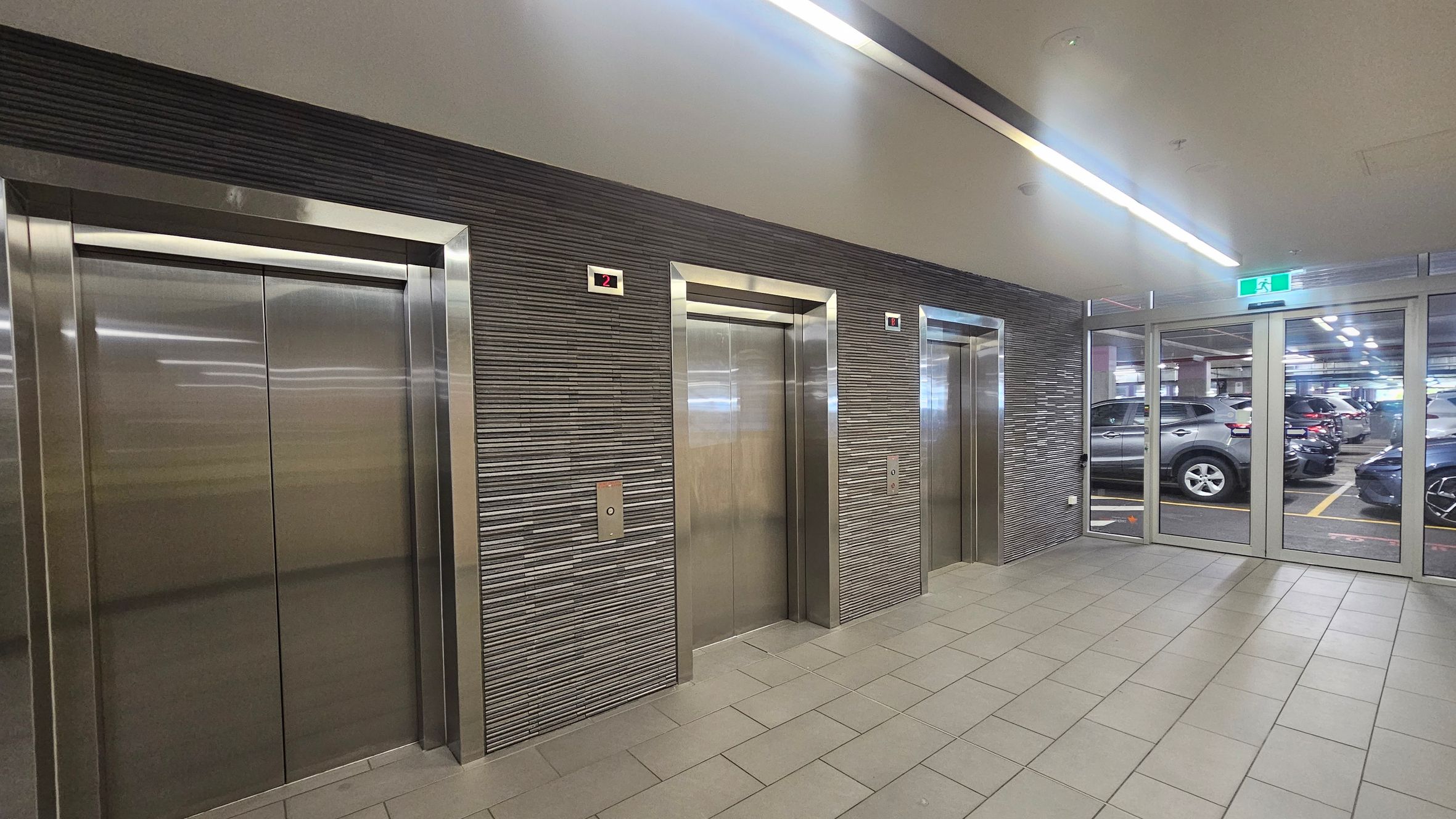Three stainless steel lift doors are set into a textured grey wall on the left and centre. Each lift has a small digital display above showing the number "2". To the right, there is a glass door leading to a car park with several parked cars visible. The floor is tiled in a light grey, and the ceiling features linear fluorescent lighting. An illuminated green exit sign is above the glass door.