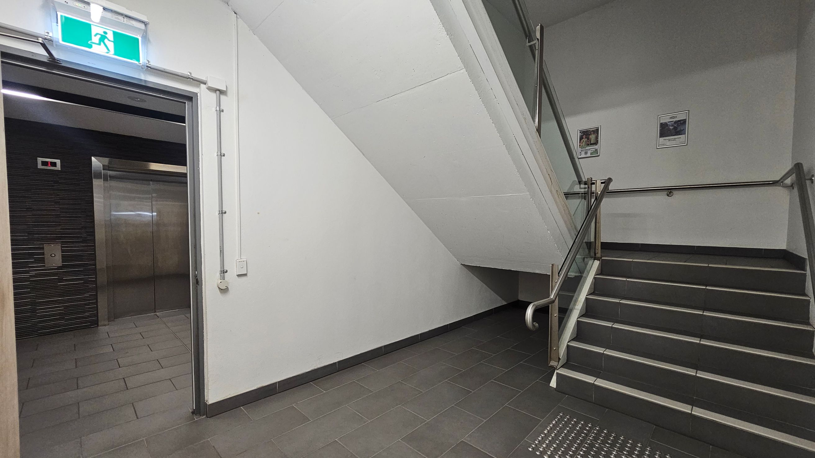 A corridor with a lift on the left, featuring metallic doors and a control panel. Above the lift is an illuminated green exit sign. On the right, a staircase with metal railings leads upwards. The walls are plain white, and two posters are mounted on the wall beside the stairs. The floor is tiled in dark grey, and the ceiling is white with visible piping.