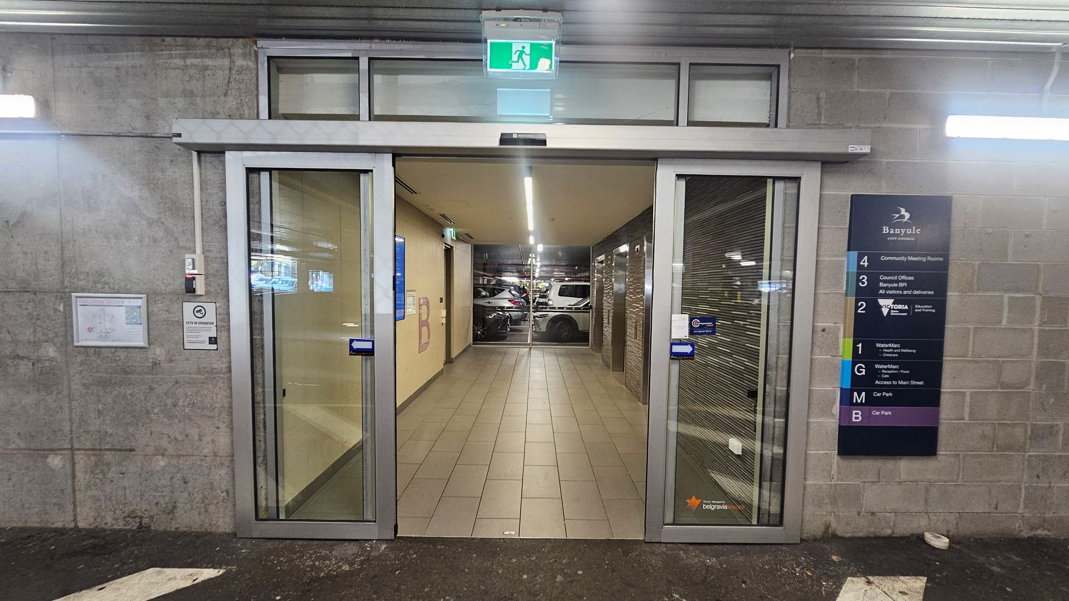 Automatic sliding glass doors lead into a corridor with tiled flooring. Above the doors, an illuminated green exit sign is visible. On the right, a directory sign lists various floors and facilities, including community meeting rooms and council offices. The left wall has a map and a sign indicating CCTV operation. Inside, the corridor is lit by overhead lights, and parked cars are visible through a glass wall at the end. The exterior wall is made of concrete blocks.