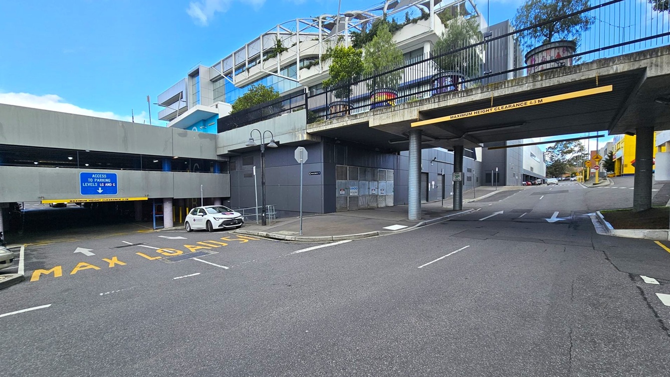 A multi-level car park entrance is on the left, with a sign indicating access to parking levels LG and G. A white car is parked nearby. The road has "MAX LOAD 15" painted on it. On the right, a road passes under a bridge with a clearance sign of 4.3 metres. Above the bridge, there are plants and a structure with a modern design. The background features more buildings and trees. The ground is asphalt, and the area is well-marked with road signs and arrows.