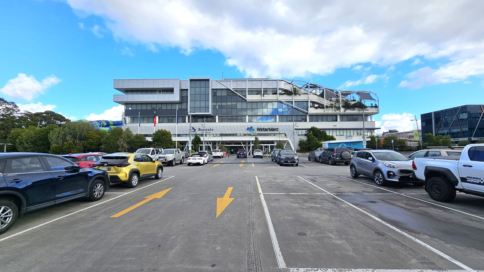 A multi-storey building with a modern design, featuring large glass windows and a white facade. The building has signs for "Banyule" and "WaterMarc" on the front. In the foreground, a car park with several vehicles, including a white convertible and a silver pickup truck. Yellow directional arrows are painted on the asphalt. Trees and flags are positioned near the entrance, and a blue structure is visible on the right side.