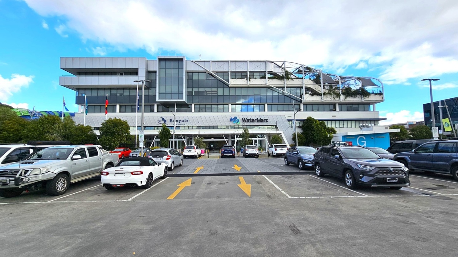 A large, modern building with a glass facade is in the centre, featuring the names "Banyule" and "WaterMarc" on the front. The foreground shows a car park with various vehicles, including a yellow car and a white truck. Yellow arrows are painted on the concrete surface, indicating traffic direction. Trees and bushes are visible on the left, and another modern building is partially visible on the right. The sky is partly cloudy.