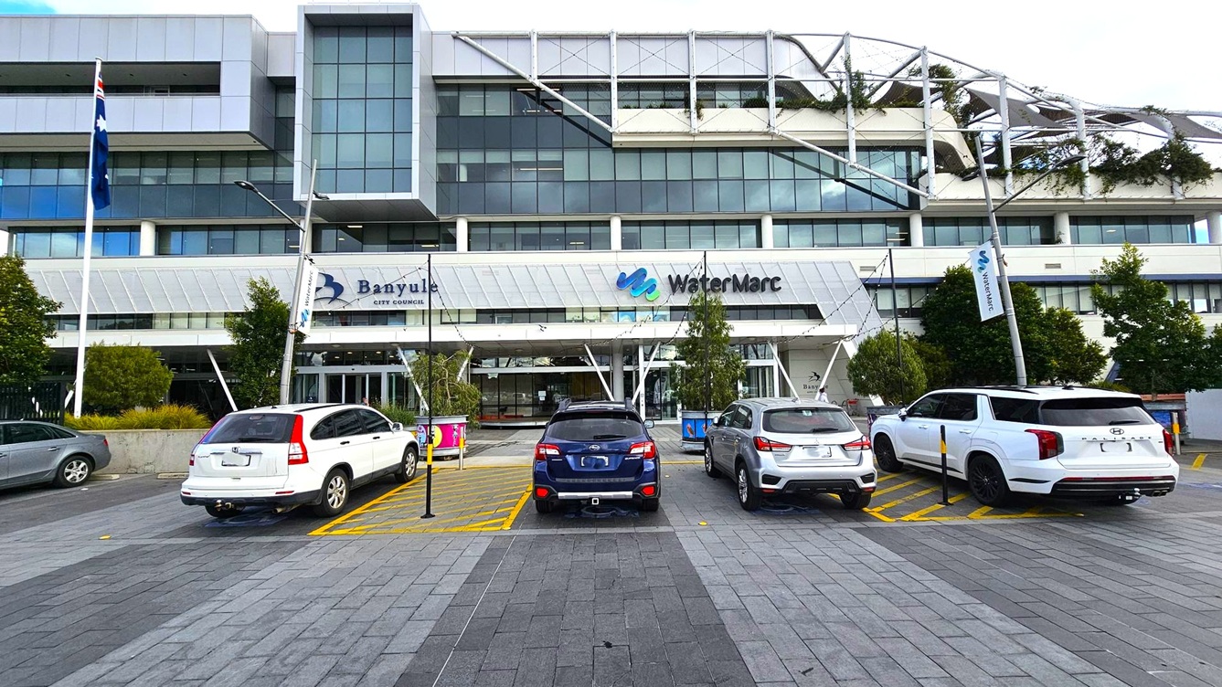 A modern multi-storey building with a glass facade and geometric design elements. In front, there are four parked cars on a paved area. The building has signs for "Banyule City Council" and "WaterMarc." There are trees and flags near the entrance. The structure features a mix of glass and metal, with greenery visible on the upper levels. The pavement is made of grey tiles, and there are banners on poles near the entrance.