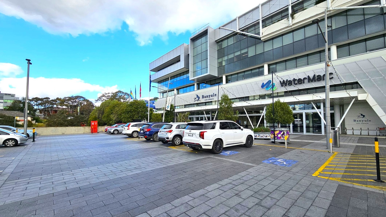 A modern building with large glass windows and a sign reading "WaterMarc" and "Banyule City Council" on the facade. Several cars are parked in the foreground on a paved area with marked parking spaces. Flags are visible near the entrance, and there are trees and bushes along the perimeter. A red utility box is positioned near the centre-left. The building has a contemporary design with angular features and a mix of metal and glass element