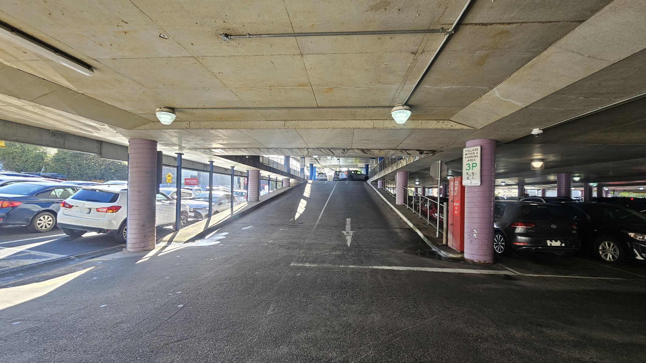 A multi-storey car park with a ramp in the centre leading upwards. On the left, several parked cars are visible, separated by a mesh fence. On the right, more parked cars are under a low ceiling with fluorescent lights. A sign on a pillar indicates parking regulations. The floor is asphalt, and the ceiling is concrete with visible pipes and lights. The area is well-lit with natural light coming from the left.