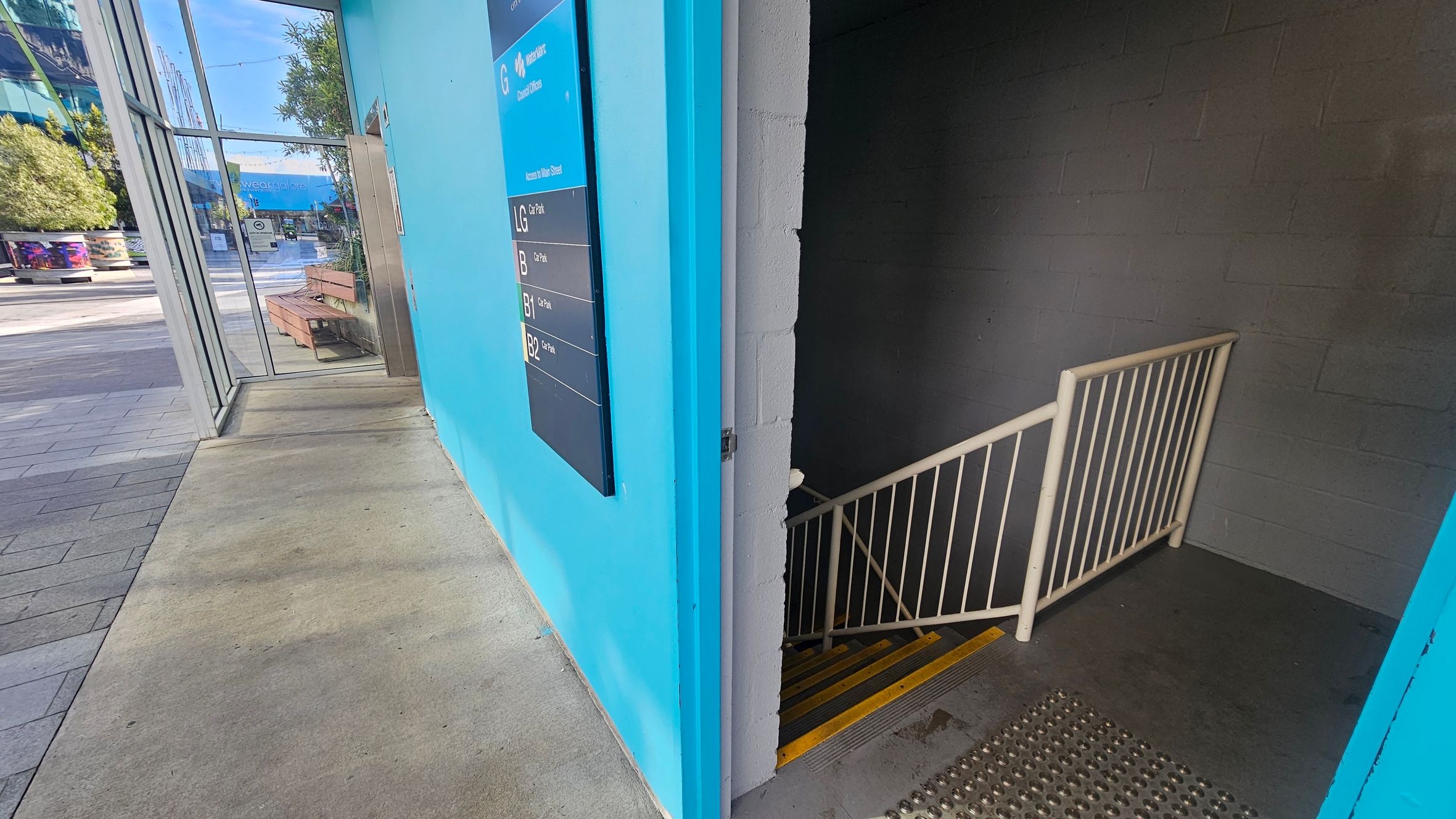 A split view with a bright blue wall on the left featuring a directory sign. Beyond the wall, there's a glass entrance with a view of an outdoor area, including benches and trees. On the right, a dimly lit stairwell with a metal railing and yellow-striped steps descends into darkness. The floor is a mix of concrete and textured metal near the stairwell entrance.