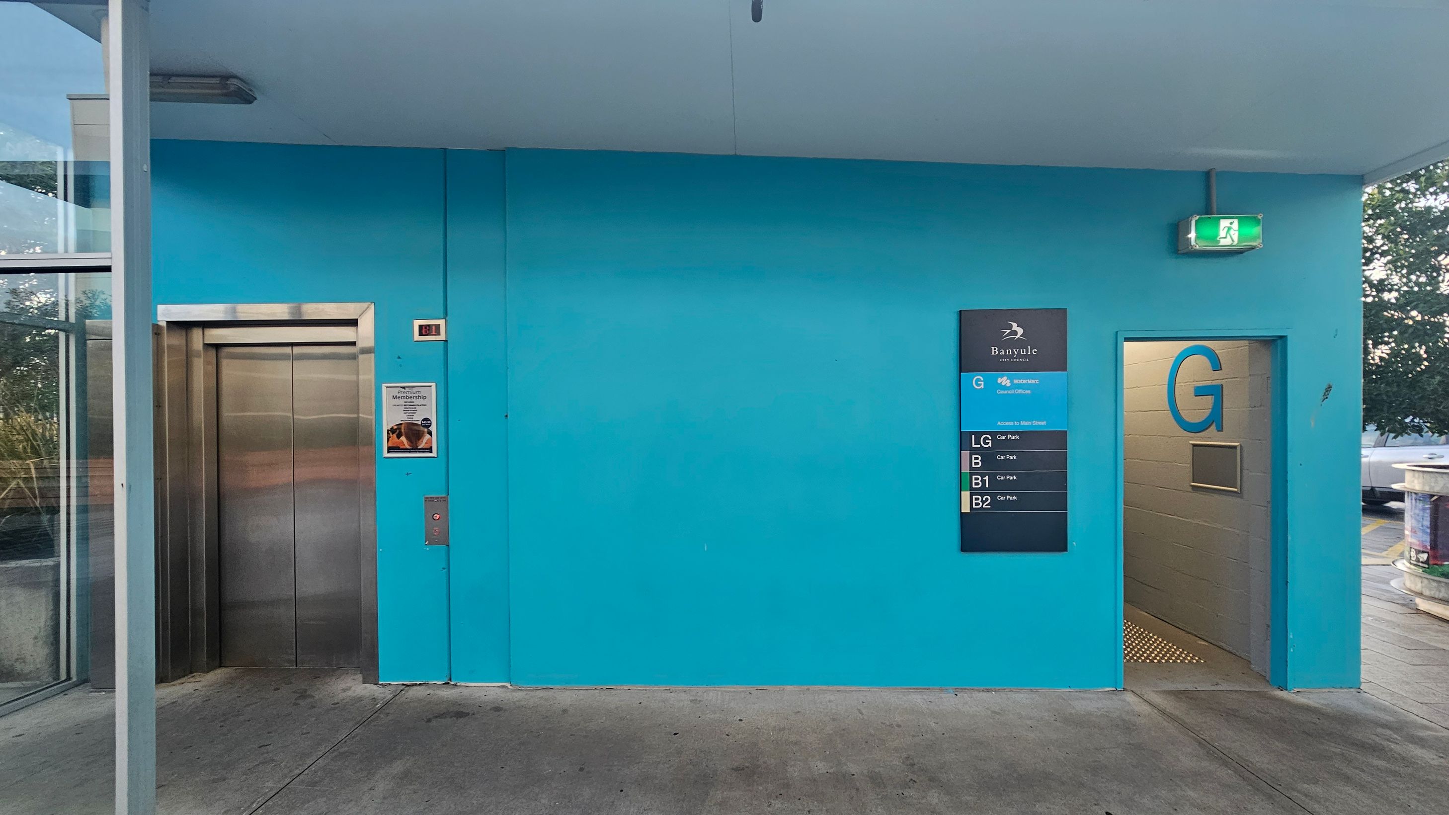 A blue wall with a lift on the left, featuring stainless steel doors. To the right, a sign displays floor information, including "G" for the ground level. An open doorway marked with a large "G" leads to a corridor. Above the doorway, an illuminated green exit sign is visible. The floor is concrete, and a glass panel is partially visible on the far left, with some greenery outside.