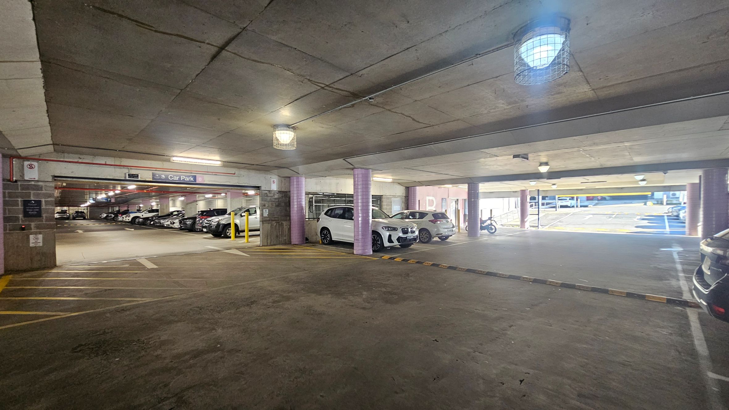 A multi-storey car park with a concrete ceiling and floor. On the left, several cars are parked in marked spaces, with a sign indicating "Car Park B". In the centre, two white cars are parked near a purple pillar. On the right, an open area leads to an exit with natural light, showing a few more vehicles and a motorcycle. The ceiling has several caged lights, and yellow lines are painted on the floor for guidance.