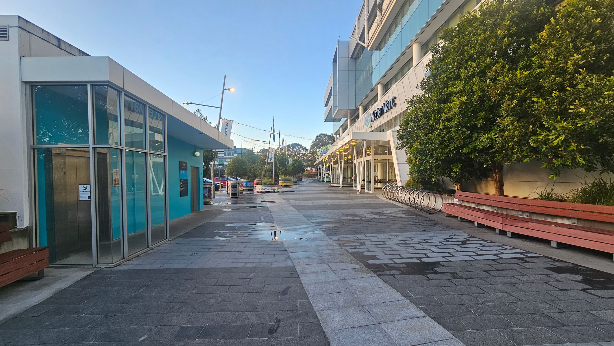 A paved walkway with a modern building on the left featuring large glass windows and a blue exterior. On the right, a larger building with a sign and a row of bicycle racks. There are trees and wooden benches along the right side. The ground is wet with puddles, and there are footprints visible on the pavement. Overhead, string lights are strung across the path. In the distance, there are more trees and streetlights.
