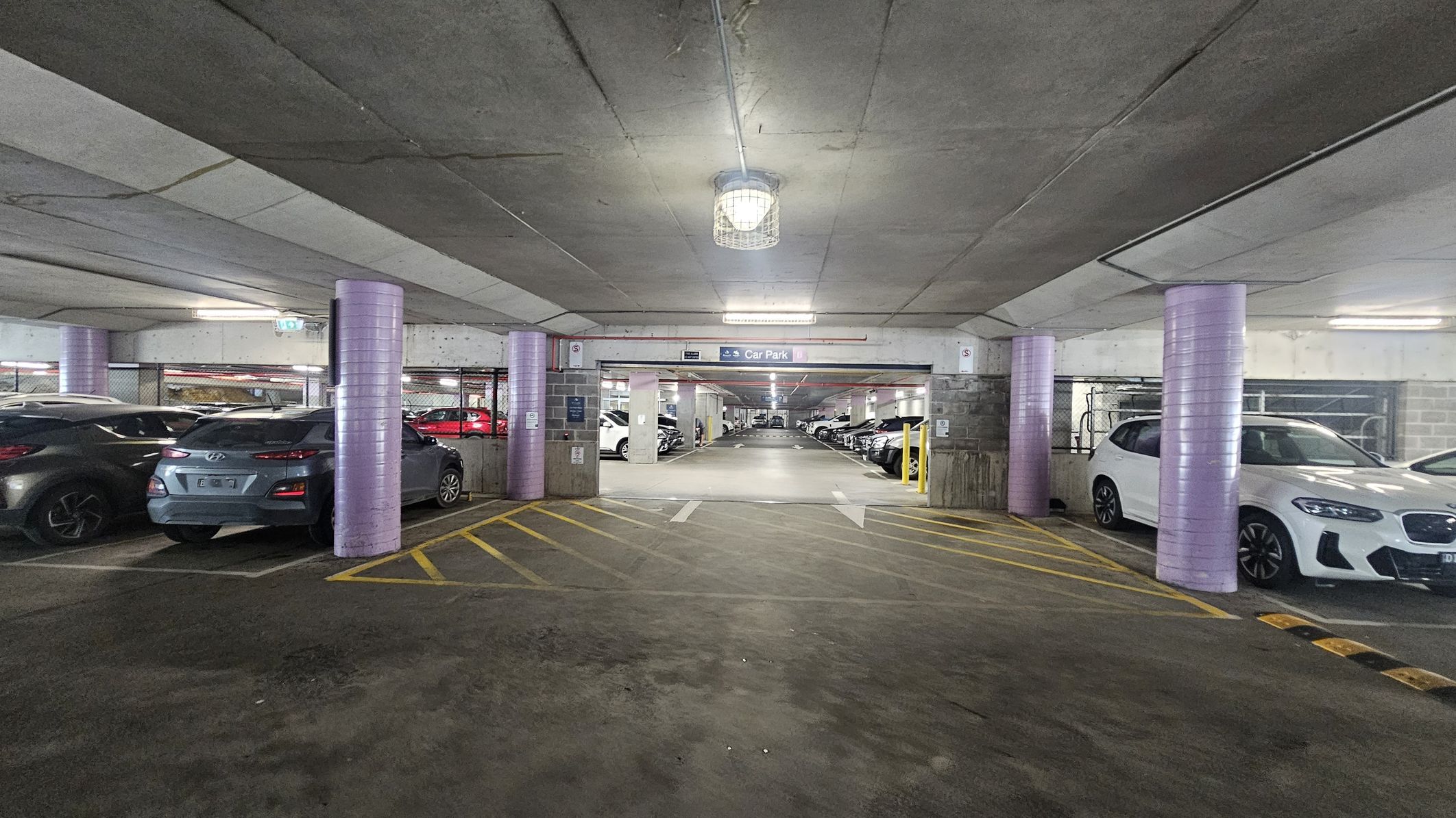 A multi-storey car park with a concrete ceiling and floor. In the foreground, there are yellow diagonal lines on the ground. The middle section shows a wide entrance to another parking area, with a sign above indicating "Car Park." The left side has parked cars, including a grey vehicle, and the right side features a white car. Purple cylindrical columns are evenly spaced throughout the area. The lighting is provided by ceiling fixtures.