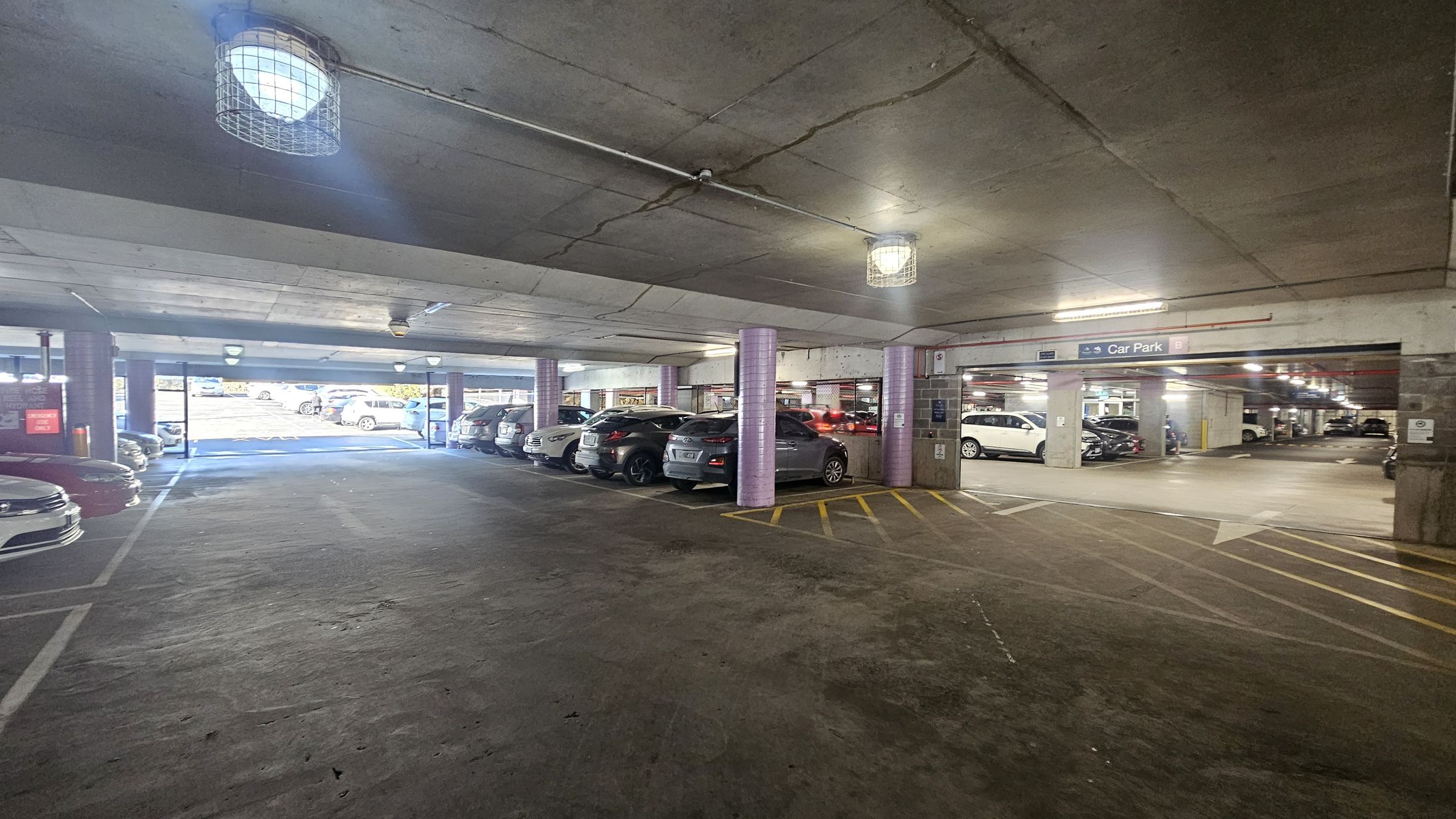 A multi-storey car park with a concrete floor and ceiling. On the left, several parked cars are visible, with a row of vehicles extending into the distance. The right side shows more parked cars and a sign indicating the car park area. The ceiling has industrial-style lights with protective cages. The floor has yellow markings and arrows for traffic direction. The background shows an exit leading to an outdoor area with natural light.