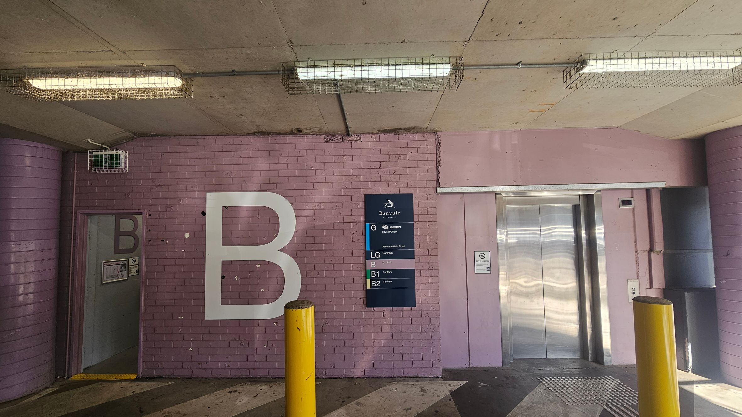 A parking garage area with a pink brick wall. On the left, a door marked with a large letter "B" leads to a stairwell. In the centre, a sign lists different levels, including "G" for ground and "B1" for basement. On the right, a silver lift with closed doors. Two yellow bollards are in front of the wall. The ceiling is concrete with fluorescent lights encased in wire cages. The floor is a mix of concrete and metal grating.