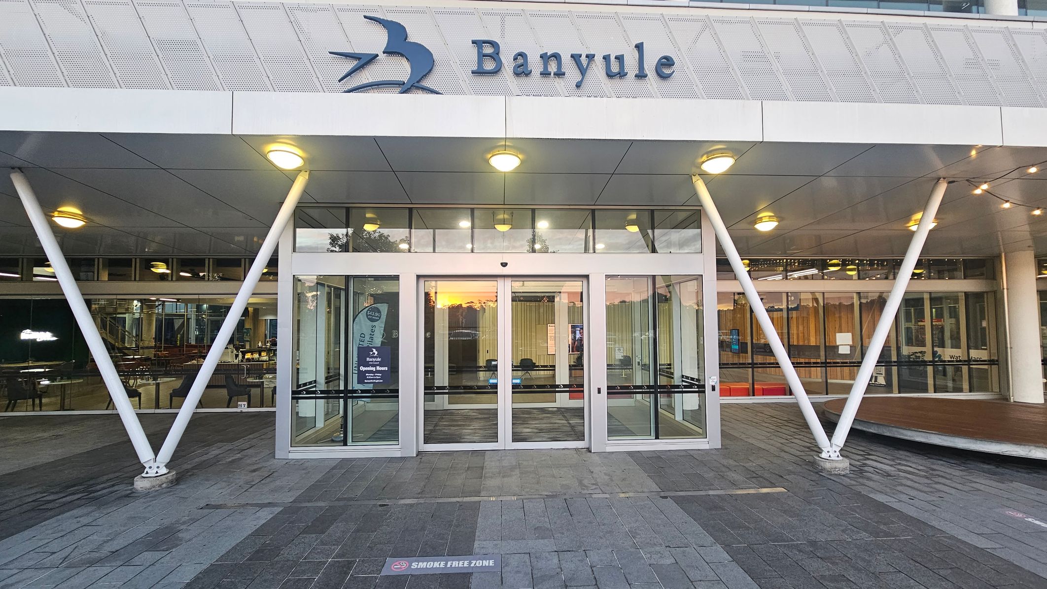 The image shows the entrance to a building with the name "Banyule" displayed above. The entrance features glass sliding doors flanked by large windows. Two white, angled support beams are positioned on either side of the doors. The ceiling is lined with circular lights. The floor is paved with grey tiles, and a sign on the ground indicates a "Smoke Free Zone." Inside, there are visible chairs and tables, suggesting a lobby or waiting area.