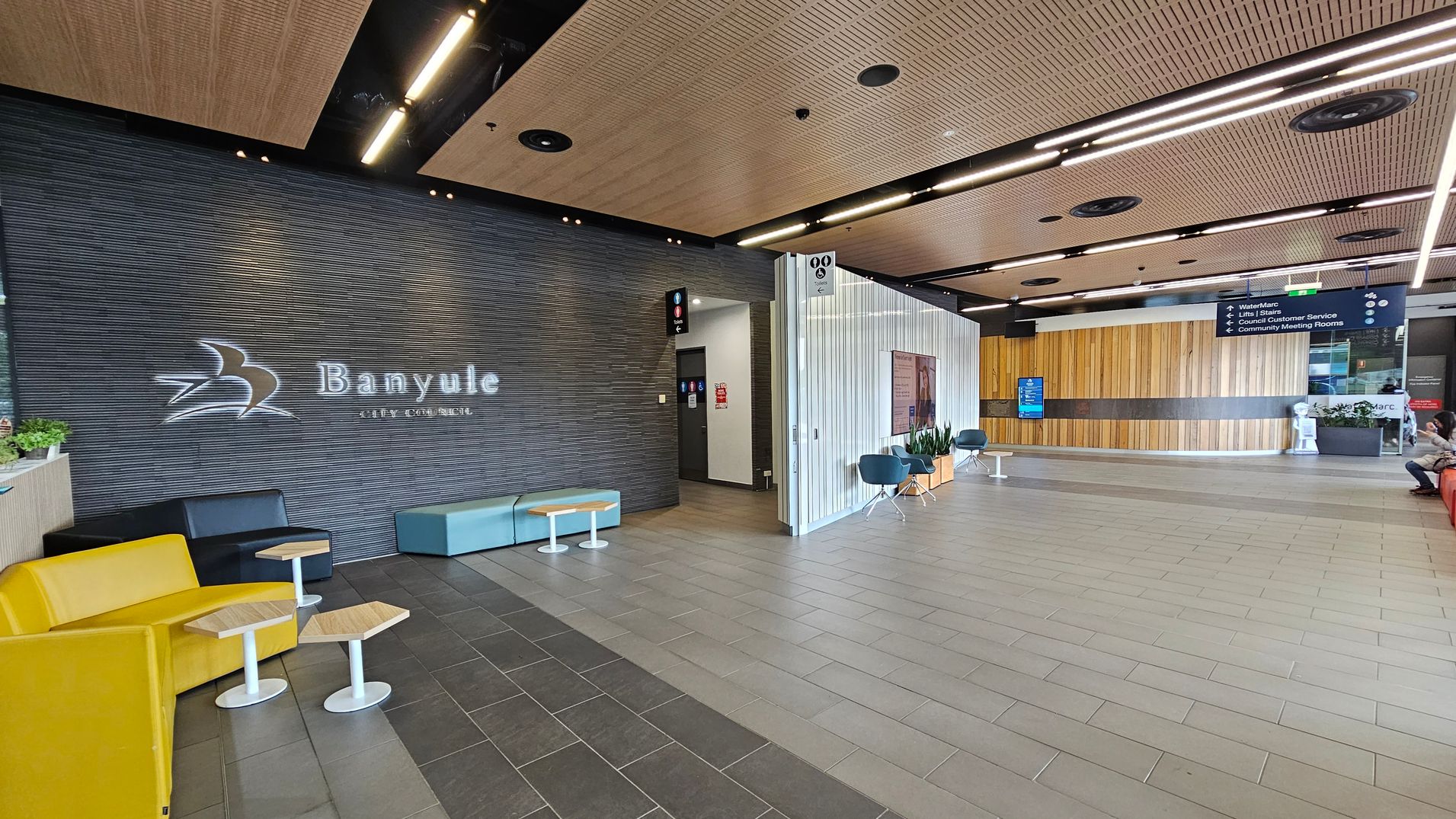 A modern interior space with a dark textured wall on the left displaying the words "Banyule City Council" and a logo. Below are yellow and black seating with small wooden tables. The floor is tiled in grey. The ceiling features wooden panels with recessed lighting. On the right, a light-coloured wall with signage and blue chairs. A wooden panelled wall is in the background, with a digital screen and a person seated on a red bench.