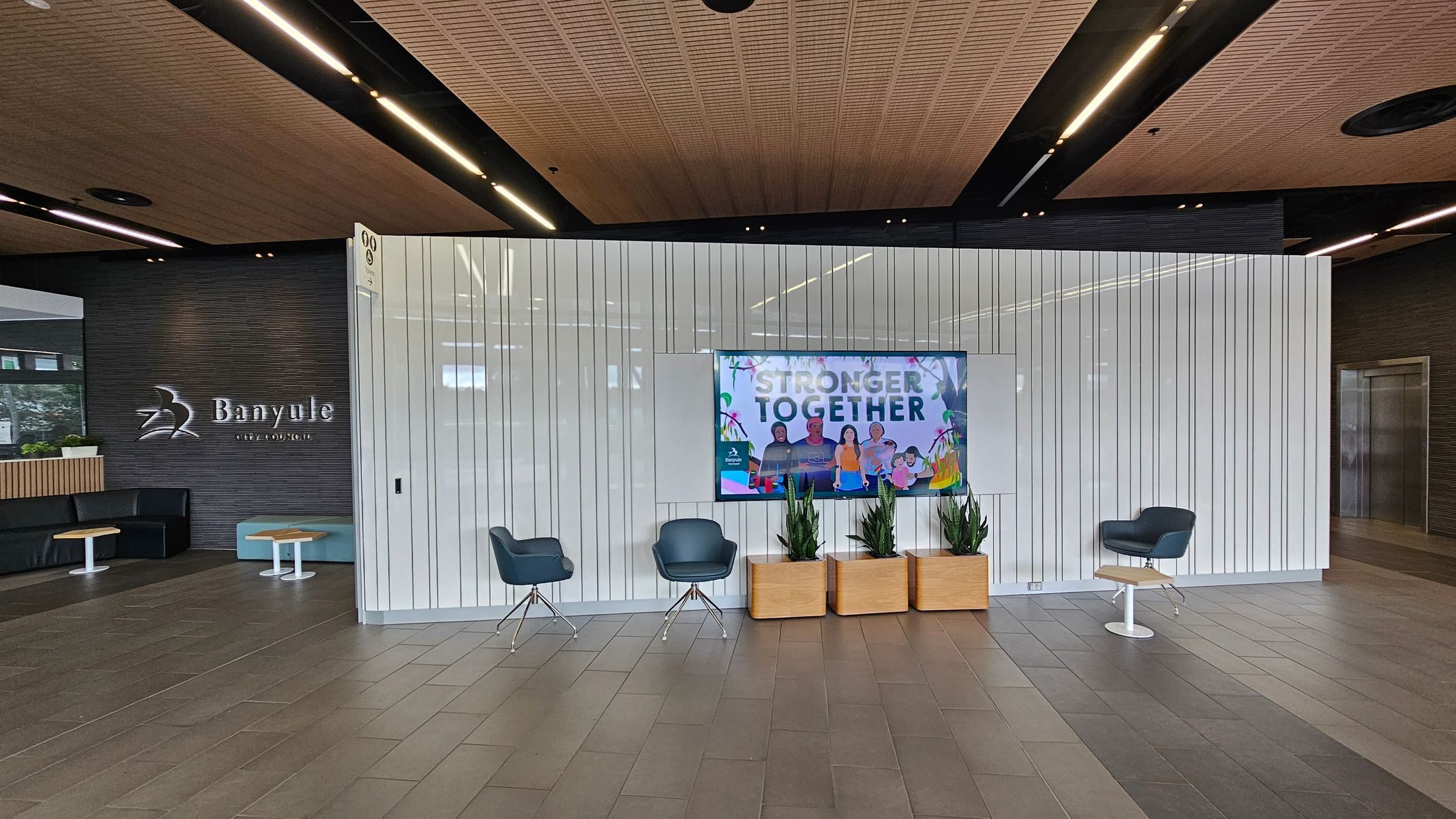 A modern interior space with a tiled floor and a wooden panelled ceiling. On the left, a dark wall features the "Banyule City Council" logo. Below it, there are black and blue seating areas with small tables. In the centre, a white panelled wall displays a screen with the text "Stronger Together" and images of people. In front of the screen, there are three potted plants. On the right, two blue chairs are positioned near a small table.