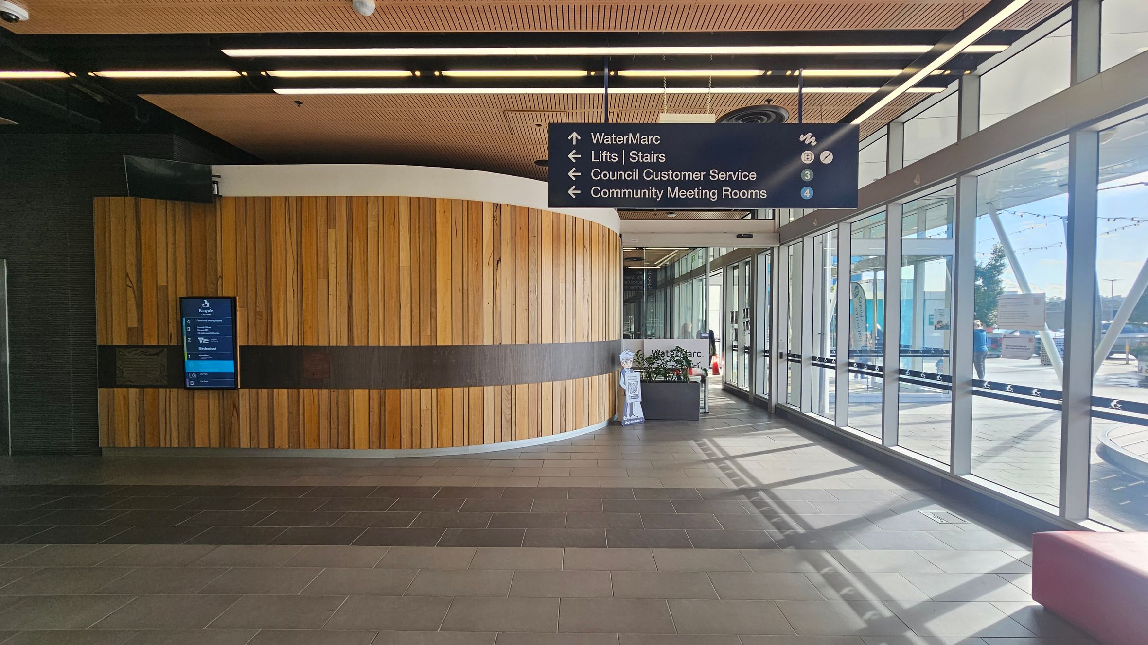 A modern interior with a curved wooden wall on the left, featuring a digital directory. Above, a sign directs to WaterMarc, lifts, stairs, council customer service, and community meeting rooms. On the right, large glass windows allow natural light, with a view of the outside. The floor is tiled in shades of grey, and the ceiling has wooden panels with strip lighting. A small reception area is visible near the windows.