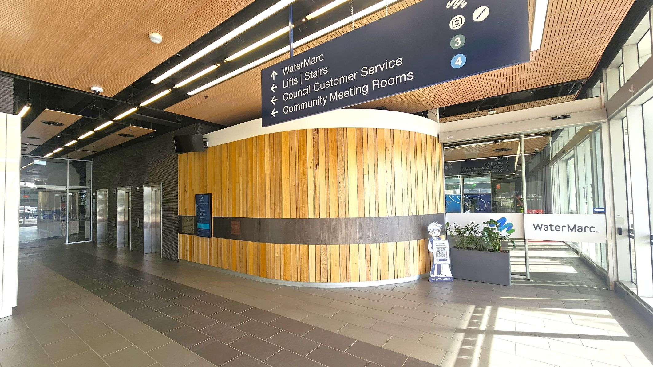 A modern interior with a wooden panelled wall on the right, featuring a digital display and a small cut-out figure holding a QR code. Above, a ceiling with wooden panels and strip lighting. On the left, there are three metallic lifts set into a dark wall. A large sign above points to various facilities, including WaterMarc and meeting rooms. The floor is tiled in shades of brown, and there are glass doors and windows on the far right with a WaterMarc sign.