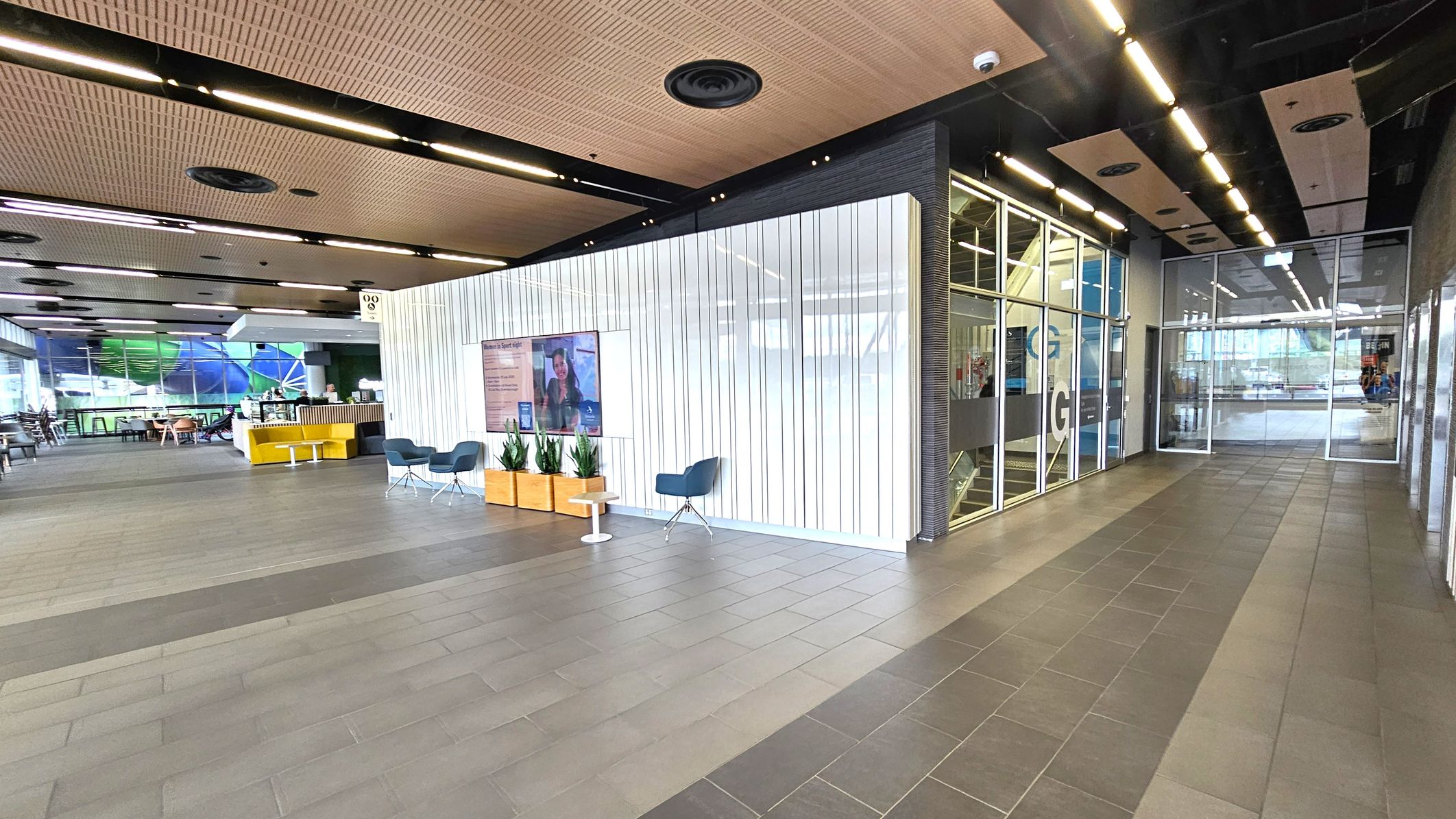 A modern interior space with a tiled floor and a wooden panelled ceiling. On the left, there is a seating area with yellow and grey chairs and a small café counter. In the centre, a white panelled wall features a digital screen and potted plants. On the right, glass doors lead to another area, with a corridor lined with glass walls. The lighting is bright, with linear ceiling lights.
