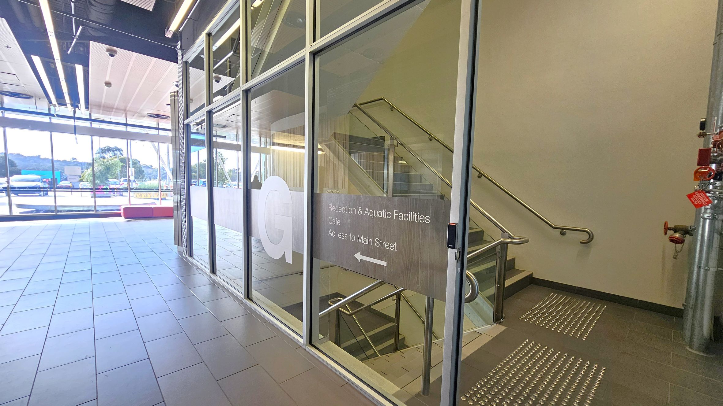 A modern interior with a glass wall on the right, featuring a door with signage indicating directions to reception, aquatic facilities, a café, and Main Street. Behind the glass, a staircase with metal railings is visible. The floor is tiled with tactile paving near the entrance. On the left, large windows allow natural light in, showing a view of parked vehicles and trees outside. The ceiling has exposed beams and lighting fixtures.
