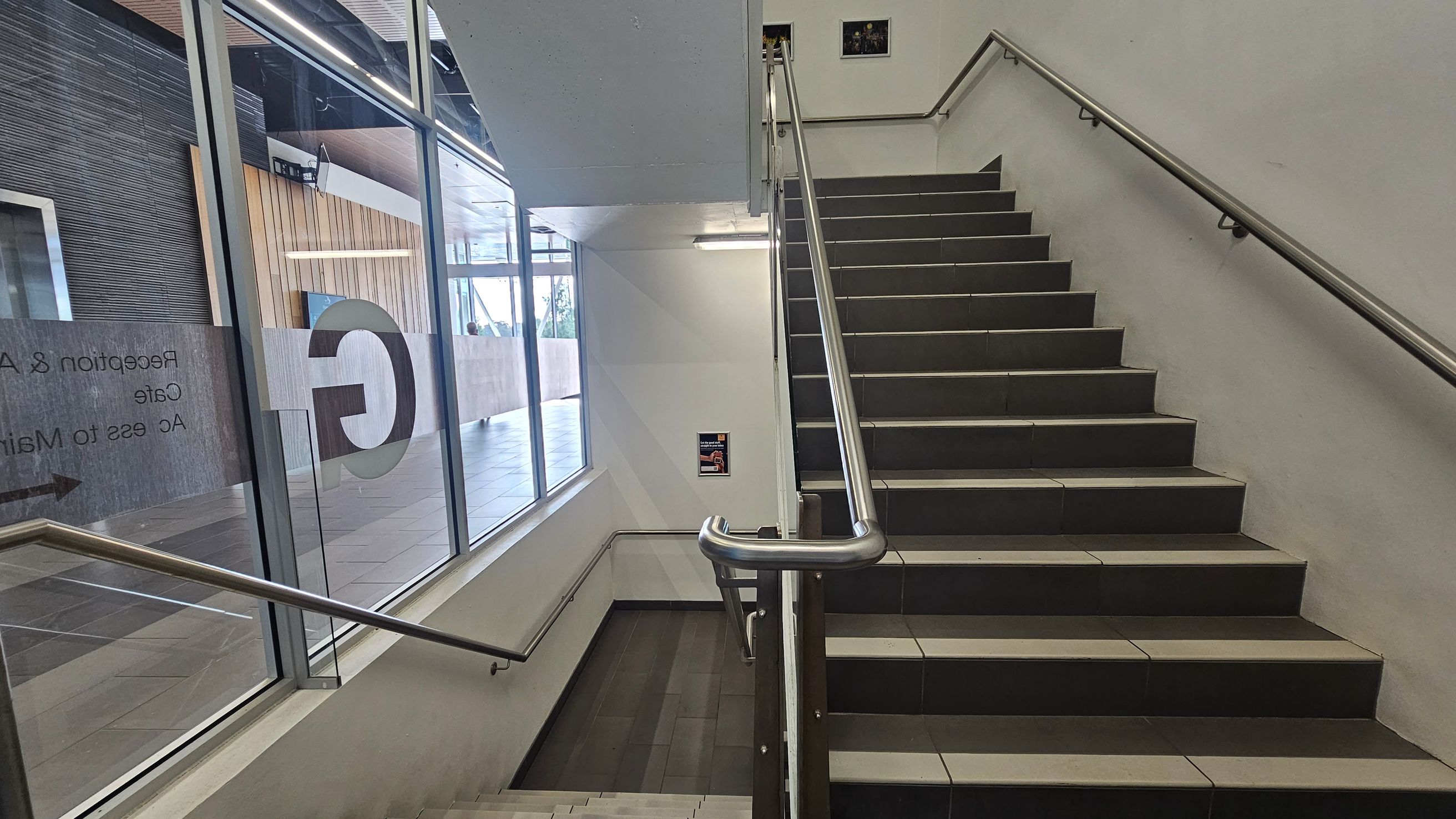 A staircase with dark steps and metal railings on both sides. On the left, a large glass window displays text and a large letter "C". Beyond the window, a tiled floor and a wooden panelled wall are visible. The ceiling is plain with a fluorescent light fixture. A small poster is mounted on the wall near the stairs.