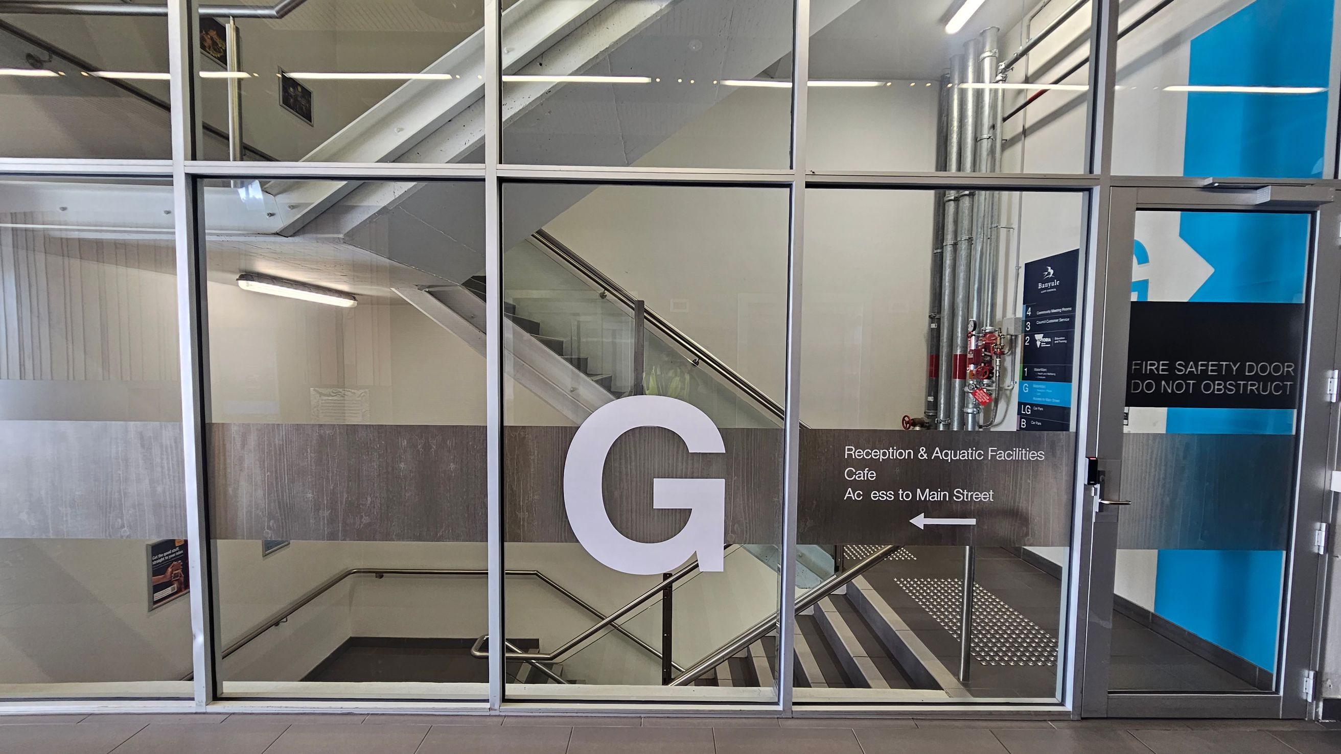 A glass wall with a large letter "G" in the centre. Behind the glass, a staircase with metal railings is visible. On the right, there are signs indicating directions to reception, aquatic facilities, a café, and access to Main Street. A blue and white sign with floor numbers is also present. Metal pipes and a fire safety door are on the right side. The floor is tiled, and fluorescent lights are on the ceiling.