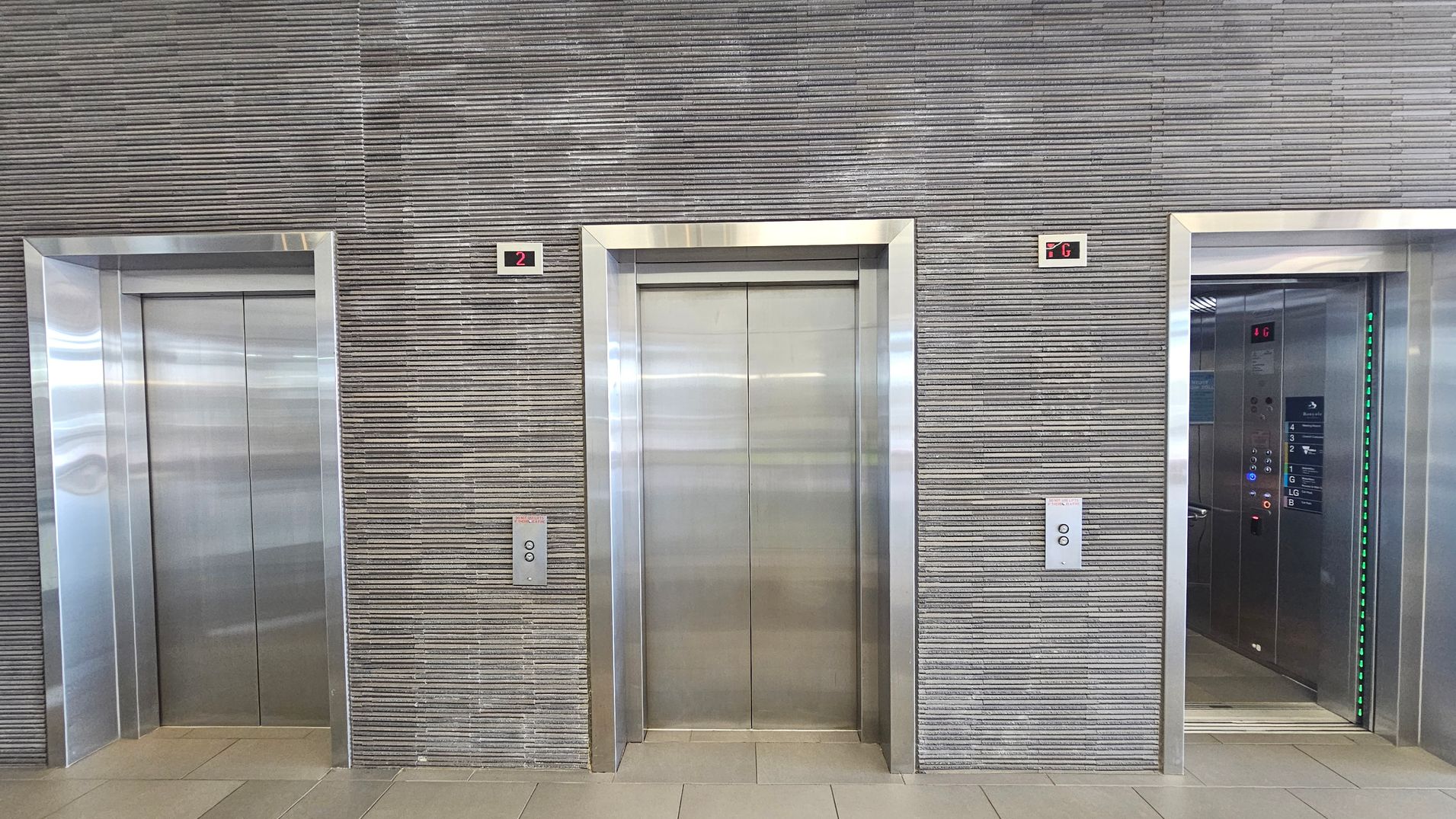 Three lift doors are set in a wall with horizontal grey lines. The left and centre lifts are closed, while the right lift is open, revealing a control panel with floor numbers and green lights. Above each lift is a digital display showing floor numbers: "2" above the left and centre lifts, and "G" above the right lift. The floor is tiled in light beige.