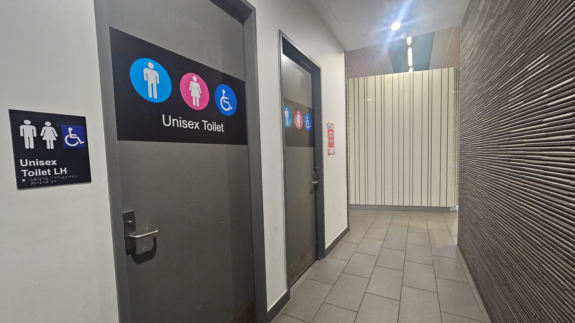 A corridor with two doors on the left, each marked as unisex toilets. The first door has a sign for a unisex toilet with left-hand accessibility, featuring male, female, and wheelchair symbols. The second door also displays unisex and accessibility symbols. The floor is tiled in a light grey, and the right wall is textured with horizontal lines. The ceiling has recessed lighting, and the far wall is covered with vertical panels.
