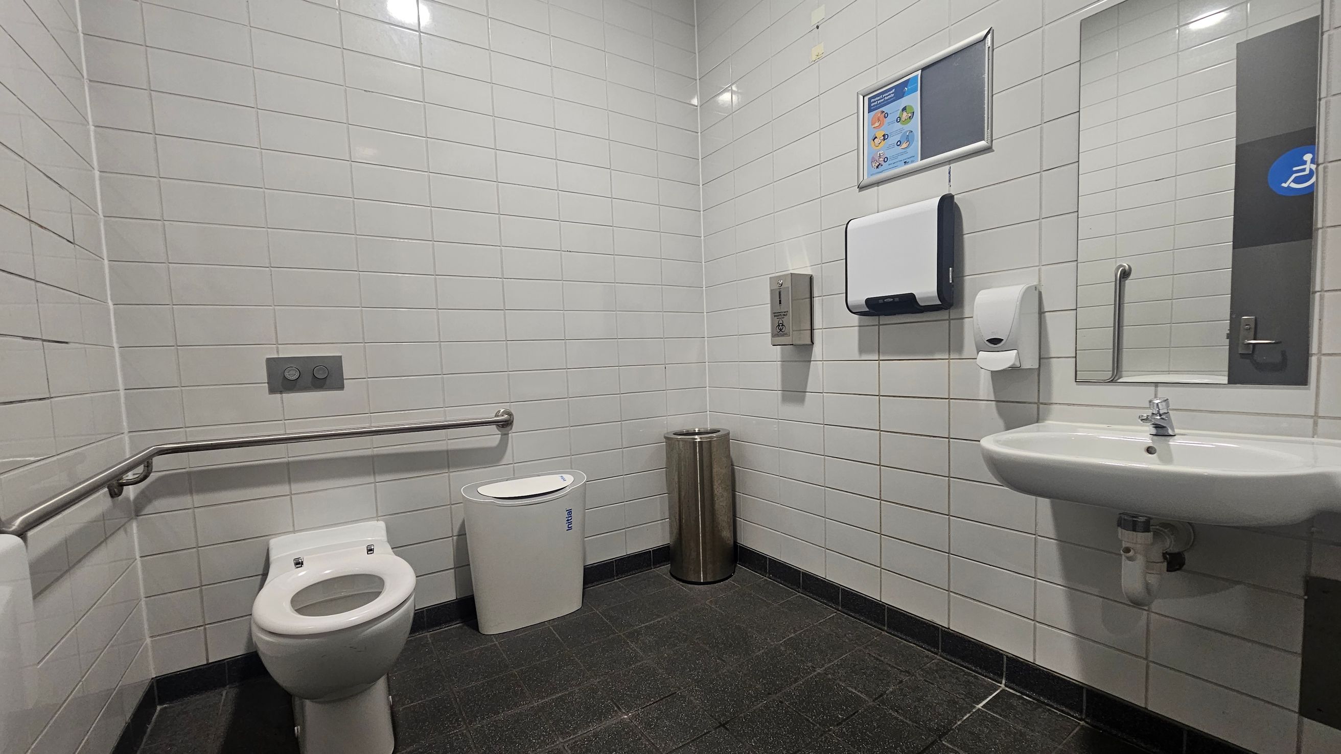 A public restroom with white tiled walls and a dark tiled floor. On the left, there is a toilet with a metal grab bar beside it. Next to the toilet is a white sanitary bin and a metallic waste bin. On the right, a wall-mounted sink with a soap dispenser above it. Above the sink, a mirror and a paper towel dispenser are visible. A sign with instructions is mounted on the wall. The ceiling has recessed lighting.