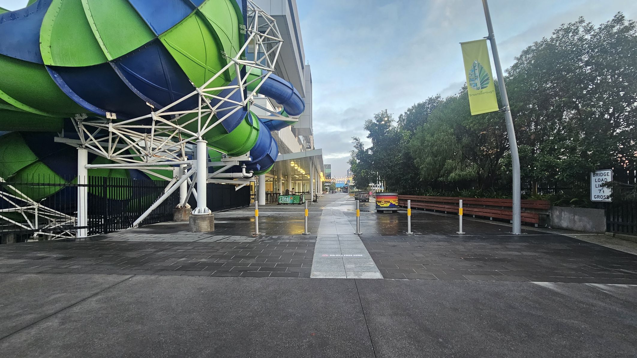 On the left, there is a large, colourful water slide structure with green and blue sections supported by white metal beams. The middle features a wet, paved walkway with several yellow-topped bollards. On the right, there are trees and a bent street lamp with a yellow banner. A sign reads "BRIDGE LOAD LIMIT 4T GROSS." The background shows a building with large windows and a distant view of more structures.
