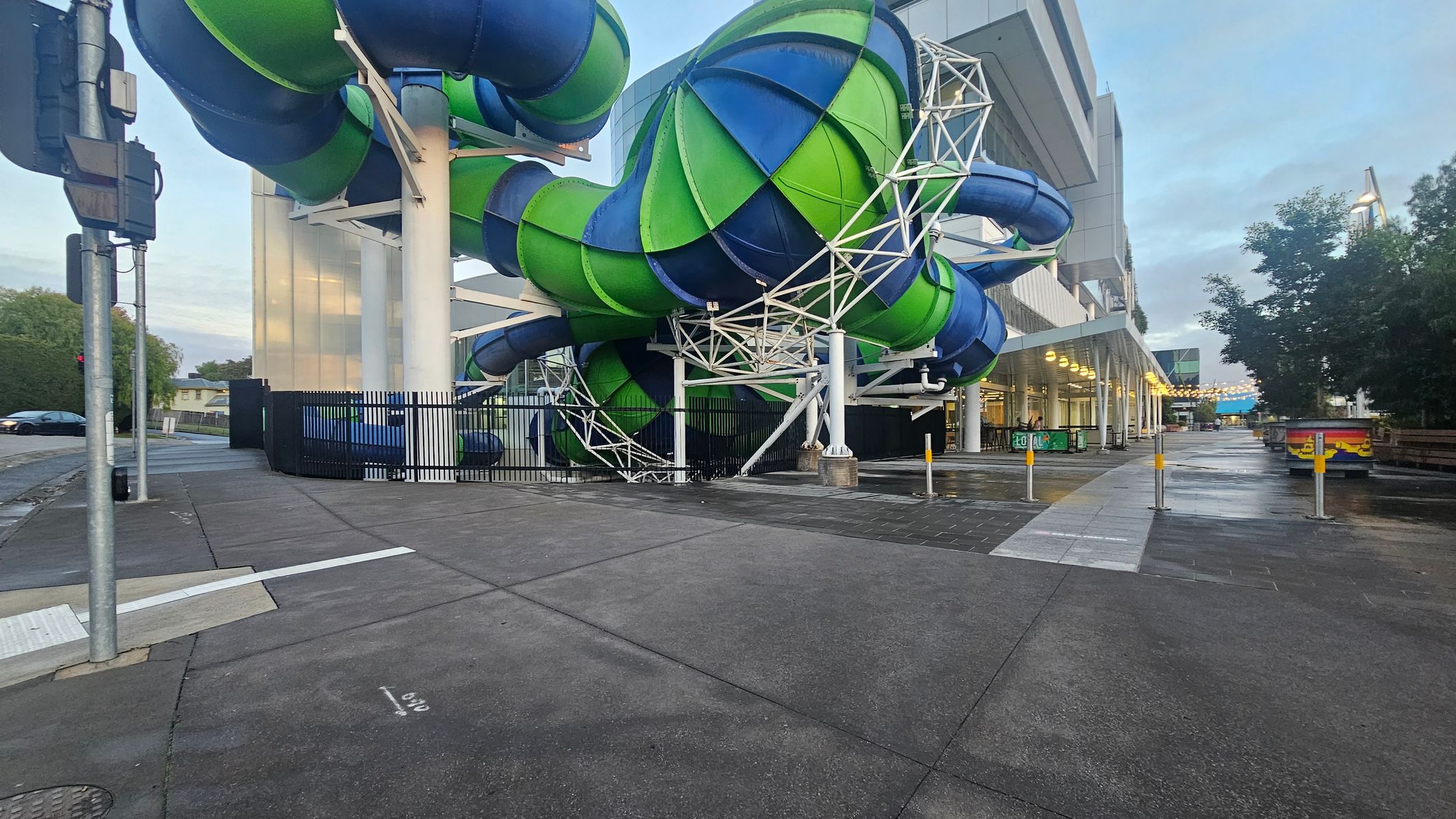 Large, intertwined water slides in green and blue dominate the centre, supported by white metal structures. On the right, a modern building with a covered walkway is lit by rows of lights. The ground is a mix of concrete and wet pavement, with bollards lining the path. On the left, a traffic light and a parked car are visible. Trees and greenery are in the background, adding contrast to the urban setting.
